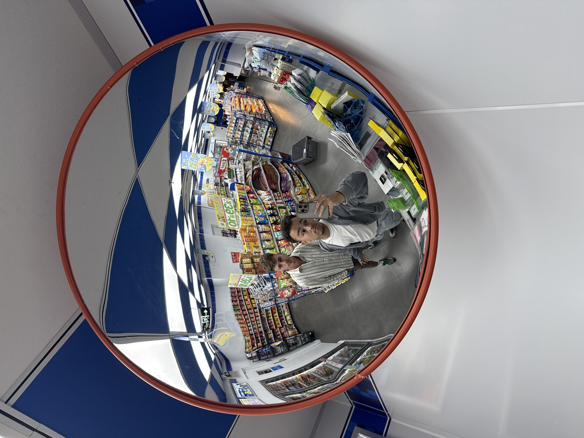 The image shows a reflection in a convex security mirror mounted in what appears to be a convenience store or small supermarket. The ceiling has a blue and white checkered pattern. Below, there are rows of shelves stocked with various food products and drinks, as well as refrigerated display cases. Two young people are visible in the reflection, positioned near the mirror. One person is holding a phone and taking a photo. The location appears to be in a city, possibly the Yuzhong District of China, judging by the context.