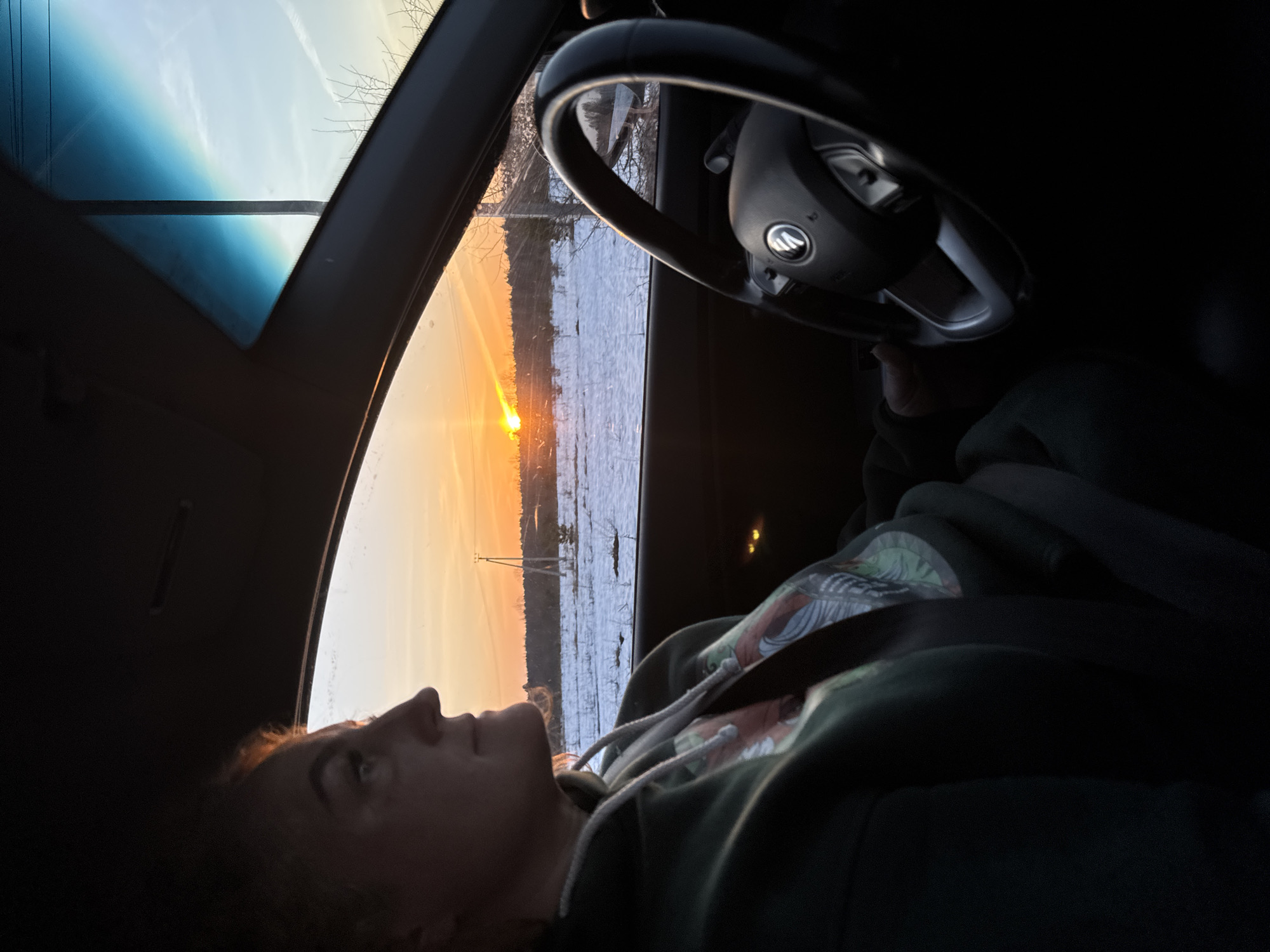A person lies reclined in the driver's seat of a car, looking out the open sunroof at a sunset over a snowy landscape. The steering wheel is visible in the foreground. A power line pole is visible on the left side of the view out the sunroof. The location cannot be confirmed as Noviy, Russia.