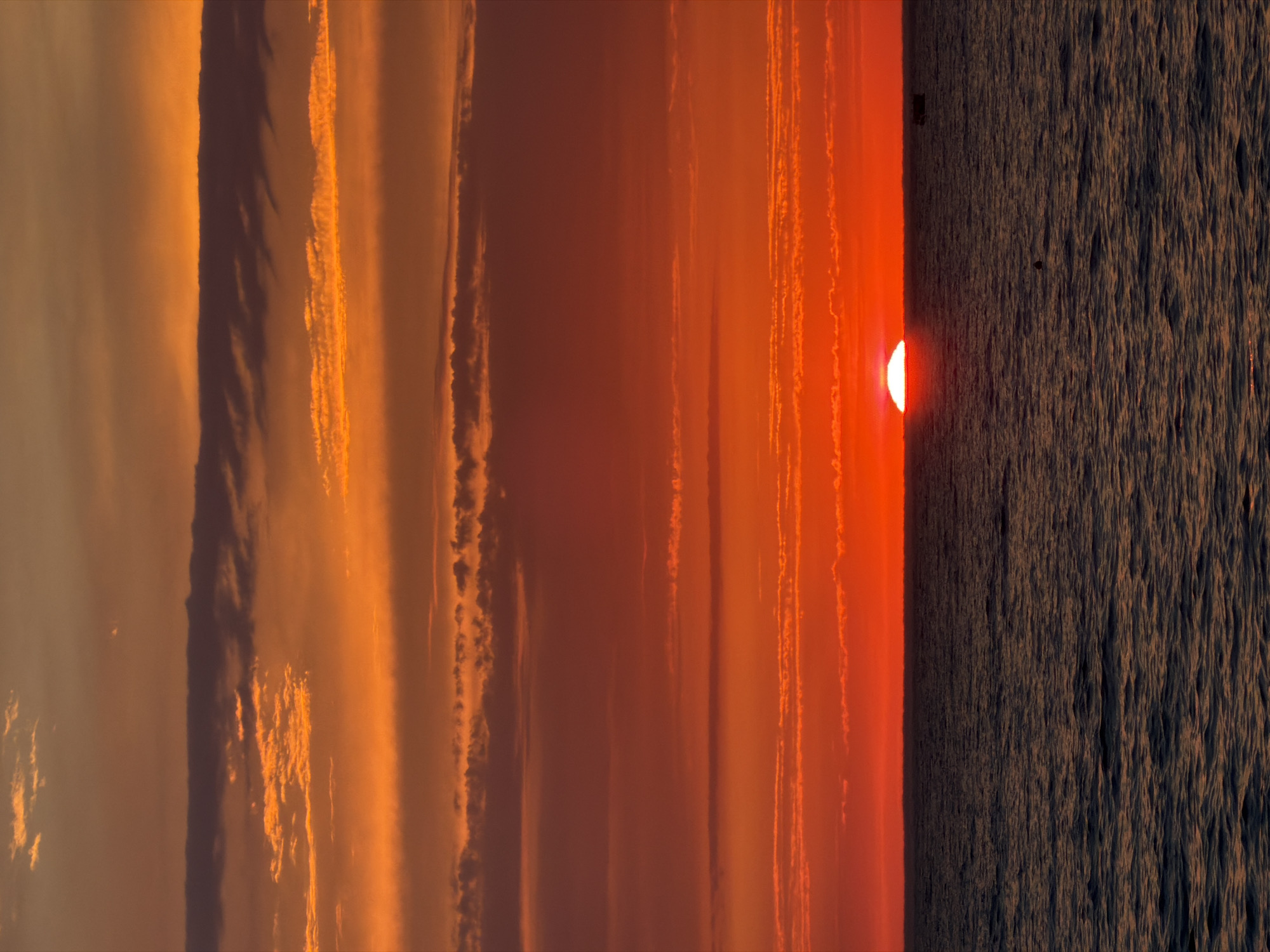 This is a photograph of a sunset over the Black Sea, likely near the city of Sochi, Russia. The sun is a bright red orb, partially submerged below the horizon, casting a vibrant orange and red glow across the sky. Numerous horizontal cloud formations are visible, varying in color from deep orange to lighter shades of red, some with darker undersides. The sea is dark and choppy, with visible waves reflecting the colors of the sky. In the distance, a small dark boat is visible on the right side of the frame, and a faint dark object, possibly another buoy or marker, is present in the middle of the frame. The overall impression is one of a dramatic and colorful end to the day.