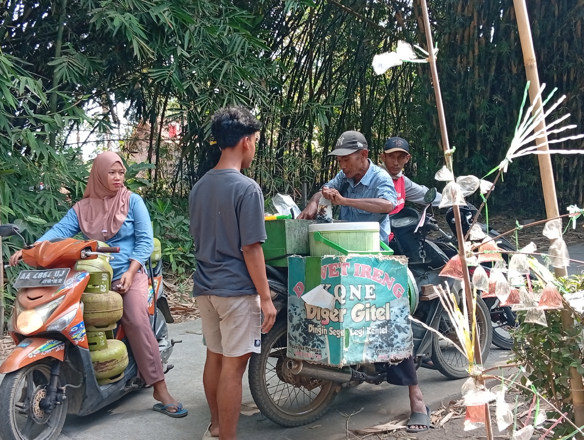In Miritpetikusan, Indonesia, on a dirt road, two men and a woman are gathered around a motorcycle laden with goods. The woman, wearing a pink headscarf and blue shirt, sits on an orange motorcycle parked to the left, holding what appears to be a gas canister. A young man in a grey t-shirt and shorts stands to her right, facing a man in a blue shirt and a black cap. This man is tending to a box attached to a motorcycle in the center, packaging items into small plastic bags. A third man, wearing a red shirt and black cap, is seated behind him. The background is dominated by dense bamboo trees, suggesting a rural or semi-rural setting. Decorative elements, possibly for a festival or event, are visible hanging from bamboo poles on the right side of the image. The motorcycle in the center displays text that appears to be "VET IENG KONE Diger Gitel" and "Terima Pesanan" along with a phone number, indicating it's a mobile vendor. The overall scene depicts a common street commerce activity in Indonesia.