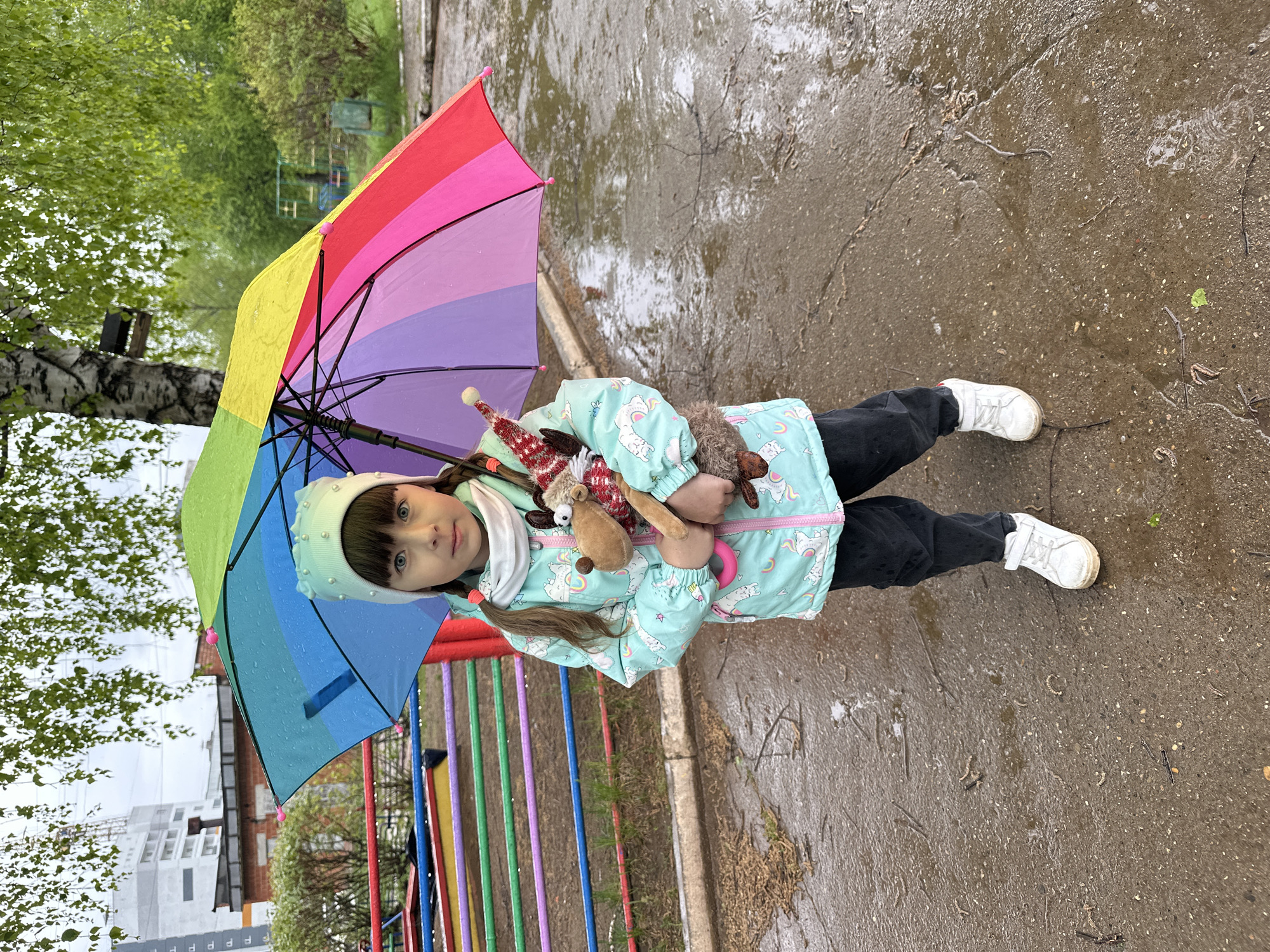 A young girl is standing on a wet sidewalk in what appears to be an outdoor area. She is holding a multi-colored rainbow umbrella and several stuffed animals. She is wearing a light blue jacket with a unicorn pattern, black pants, and white shoes. A light-colored beanie is on her head. The background includes trees, a colorful play structure, and a gray building in the distance. Based on the wet pavement, it has recently rained. The location is Chaikovsky, Russia.