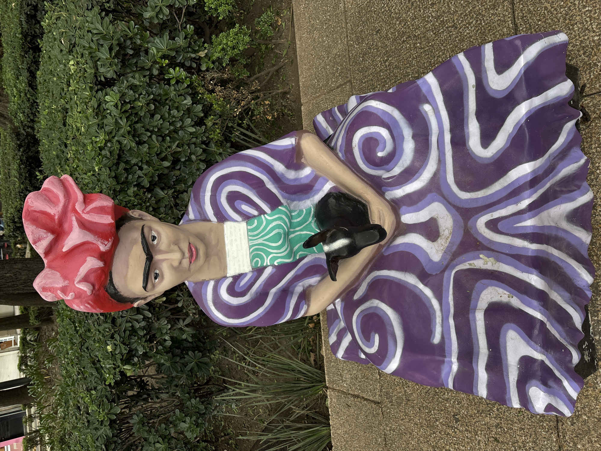 A statue of Frida Kahlo is seated outdoors, holding a small black dog. She is wearing a purple and white swirl-patterned dress with a green and white patterned bib. Her hair is styled in a large red bow on top of her head. The statue is on a paved surface in front of a dense green hedge. The statue is located in the Frida Kahlo Museum (Casa Azul) in Coyoacán, Mexico City, Mexico.