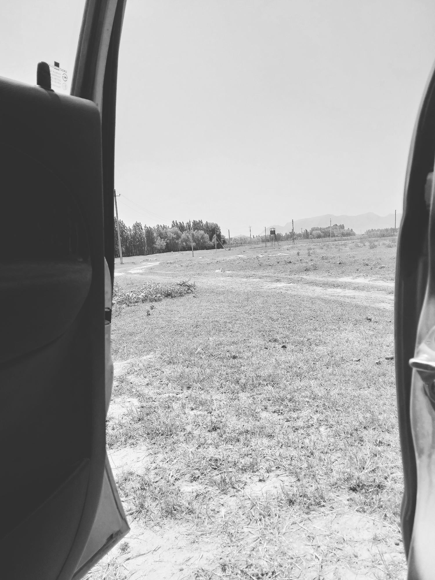 The image captures a view through an open car door, looking out onto a rural landscape in Aravan, Kyrgyzstan. The foreground is dominated by the dark interior of the car, specifically the car door panel on the left and a portion of the car's frame on the right. Beyond the car, a dry, grassy field stretches into the distance. A dirt path winds through the field, leading towards a line of trees in the mid-ground. Scattered throughout the field are utility poles with wires, and a watchtower-like structure can be seen in the distance. The background consists of rolling hills and mountains, shrouded in a light haze. The sky is bright and overcast. The overall impression is one of a remote, open, and somewhat desolate rural setting.