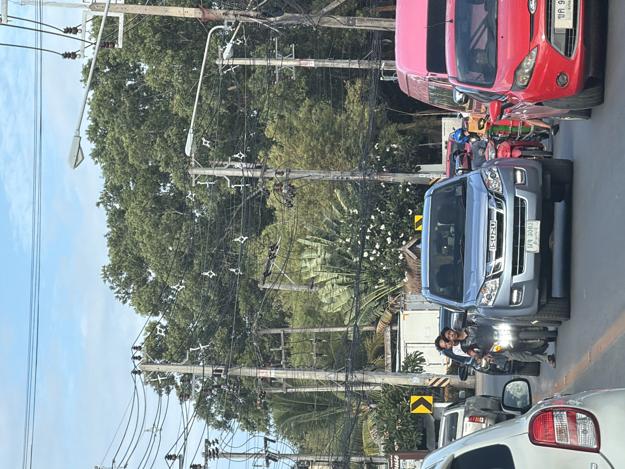 This is a daylight, eye-level, medium shot captured from a vehicle in traffic on a street in the city of Ko Samui District, Thailand. In the foreground, the left side of a white car is visible, followed by a silver SUV, and then a red Ford Fiesta. A pink van is visible between the red car and the silver SUV. In the center of the frame, two young men are riding a motorcycle, with the rider looking directly at the camera. The street is congested with vehicles. Overhead, a complex network of electrical wires hangs from utility poles, with a bright blue sky visible above. Lush green trees and foliage line the street, with a few buildings and signs also present. The overall atmosphere is that of a typical busy street scene in Thailand.