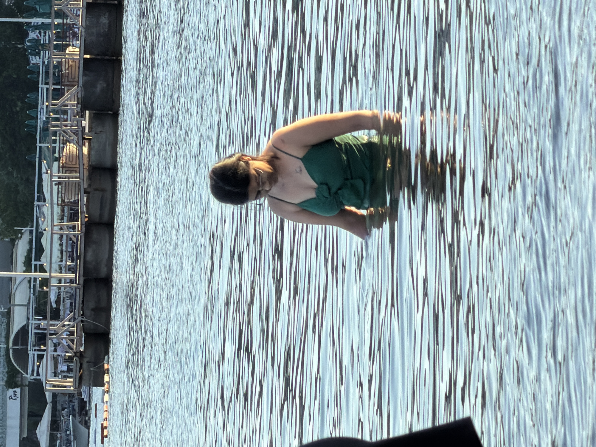 A woman in a green swimsuit is wading in the water near a concrete pier. The pier is situated near outdoor restaurant seating with umbrellas and metal frames. The location appears to be a body of water in Budva, Montenegro.