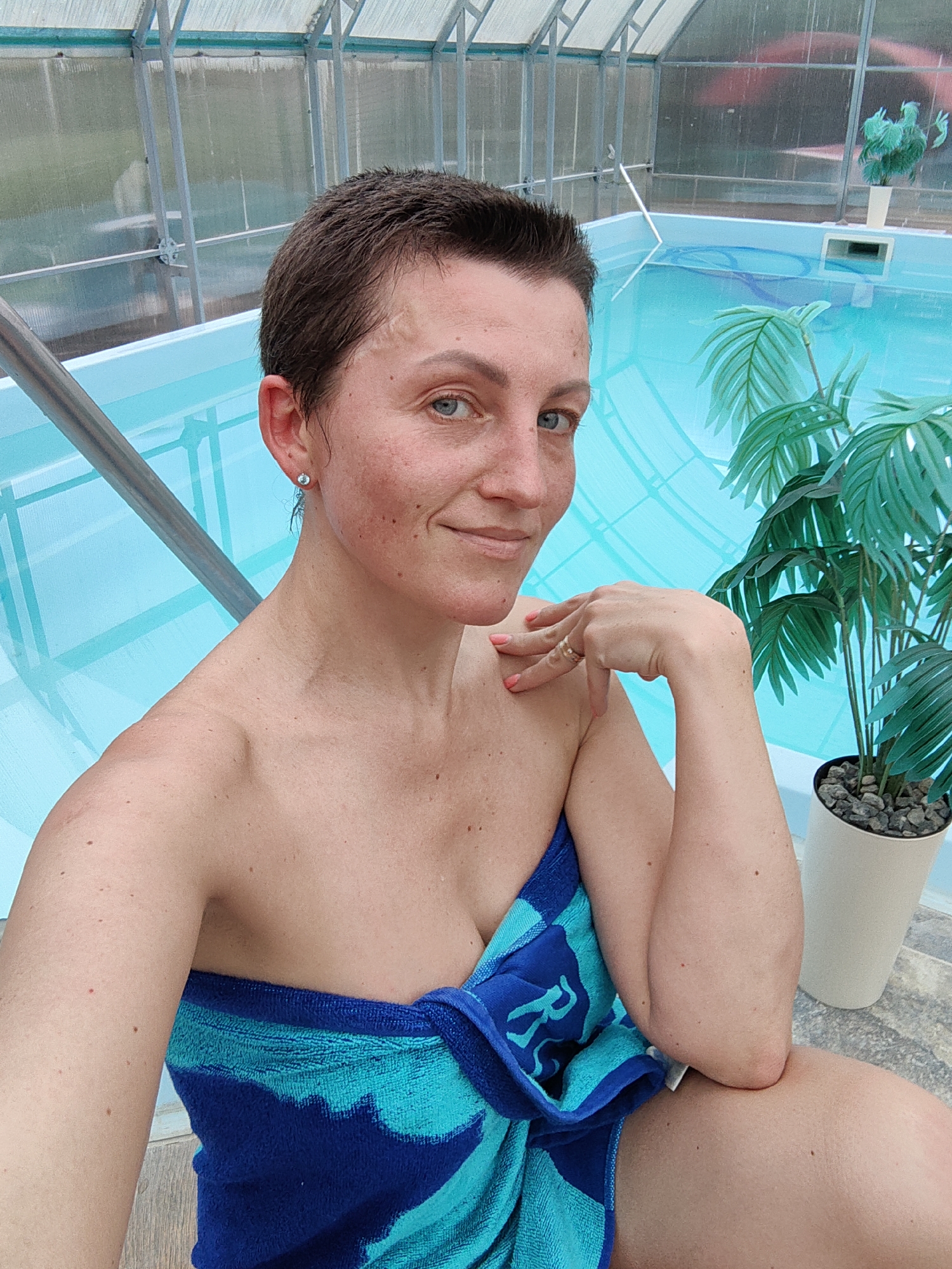 The image shows a woman in a blue and turquoise striped towel, seated next to an indoor swimming pool. The pool is filled with clear blue water and surrounded by a tiled floor. The background features a greenhouse-like structure with a transparent roof and metallic supports. Artificial palm plants in white pots are visible near the pool. The woman is looking at the camera with a slight smile. Her short brown hair is wet, and she is wearing a small earring. Her right arm is resting on her chest, with her hand near her shoulder. A ring is visible on her finger. The scene appears to be a recreational facility in the Фаніпальскі сельскі Савет, Belarus.