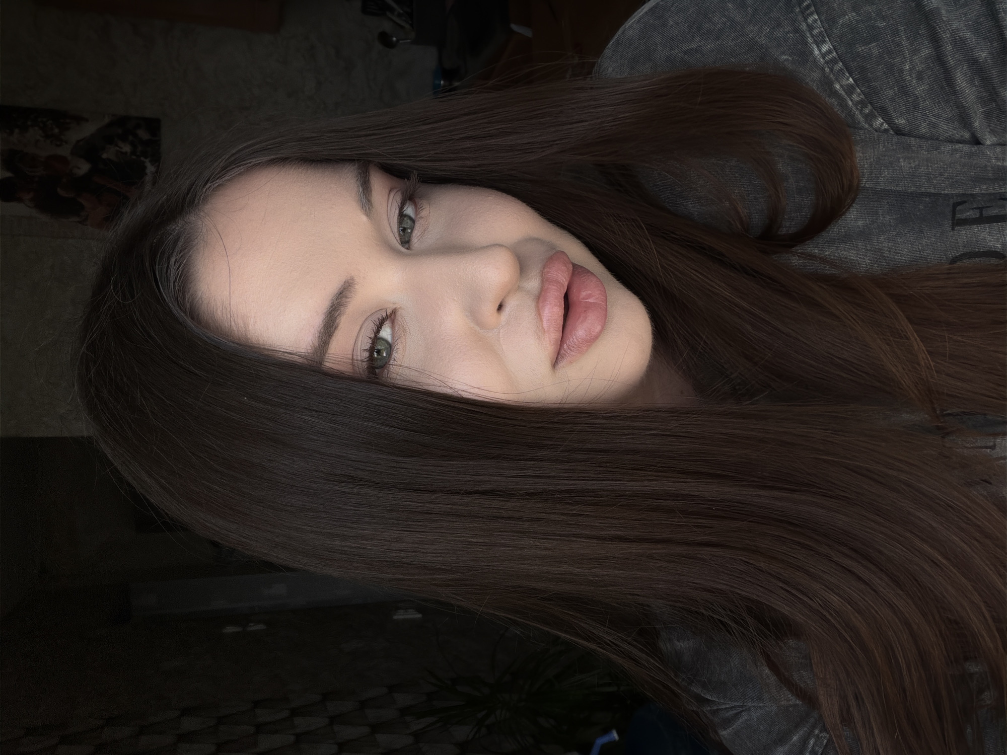 The image is a close-up, indoor, eye-level, selfie-style portrait of a young adult woman with long, dark brown hair. Her hair is parted on the left side and falls down around her face and shoulders. She is wearing a dark gray, textured t-shirt. Her skin appears fair with light makeup, including eyeliner, mascara, and lip gloss. She is looking slightly to her right, with her head tilted. The background is dimly lit and appears to be a room, with some textured wallpaper on the left and a blurred background on the right that suggests furniture or shelves. A framed picture is visible on the wall in the upper right portion of the image. The overall lighting is soft, with the primary light source coming from the front, illuminating her face. The image quality is good, with clear details of her features.