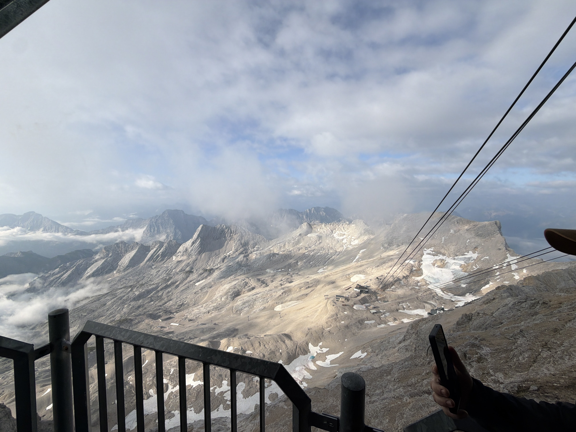 The image depicts a panoramic view of a mountainous landscape, likely taken from an elevated platform or cable car station. In the foreground, a black metal railing with vertical bars and a rounded cap on a post suggests a viewing area. To the right, a person's arm and hand are visible, holding a black smartphone. The person is wearing a dark jacket and a light-colored cap.
The main subject is the expansive mountain range stretching into the distance. Jagged peaks, some with patches of snow and ice, dominate the scene. Clouds drift through the valleys and around the mountains, creating a dramatic and misty atmosphere. The terrain appears rugged and barren, with shades of gray and brown rocks, and occasional lighter patches of snow. Several structures, possibly buildings or ski lifts, are scattered across the slopes in the middle ground.
Several thick, dark cables, likely part of a cable car system, extend diagonally across the upper right portion of the image. The sky above is partly cloudy, with patches of blue visible between the white and gray clouds. The lighting suggests it is either early morning or late afternoon, casting shadows and highlighting the contours of the mountains.
Given the context of Grainau, Germany, this view is almost certainly of the Zugspitze, Germany's highest peak, and its surrounding Alpine environment. The cable car system and the viewing platform are characteristic of the infrastructure developed for tourism and access to the summit.