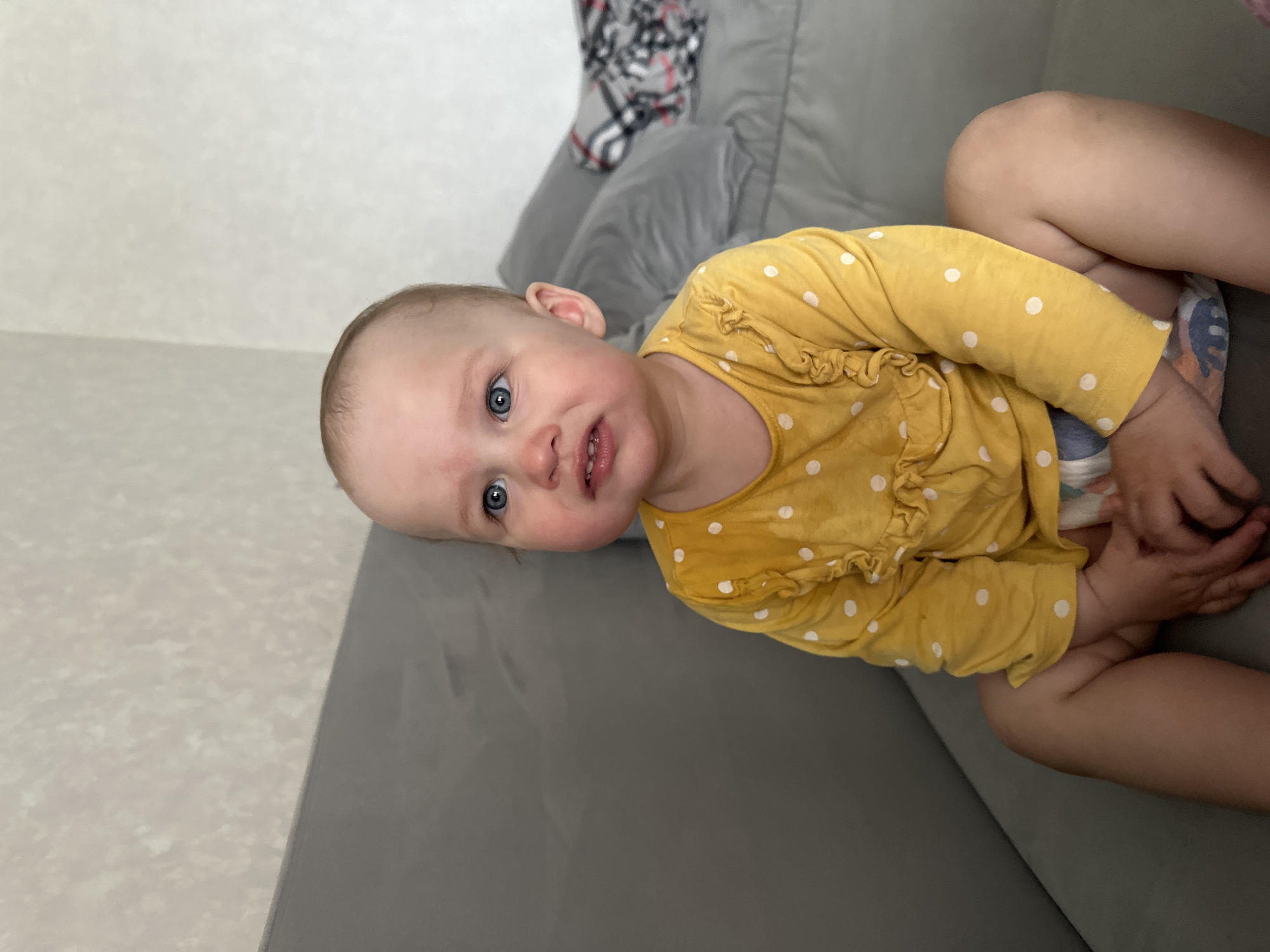 A young child is lying on a grey upholstered sofa indoors, looking directly at the camera with a curious expression. The child has fair skin, blue eyes, and light-colored hair, and their mouth is slightly open. They are wearing a long-sleeved yellow shirt with white polka dots and ruffles on the front. A patterned diaper is visible on the child's bent leg.
The background shows a light-colored wall with a subtle texture, and another section of the grey sofa, where a plaid blanket or throw with grey, white, black, and red patterns is draped. The lighting appears soft and even, suggesting a daytime setting. There is no visible text in the image. The scene is located in Chaikovsky, Russia.