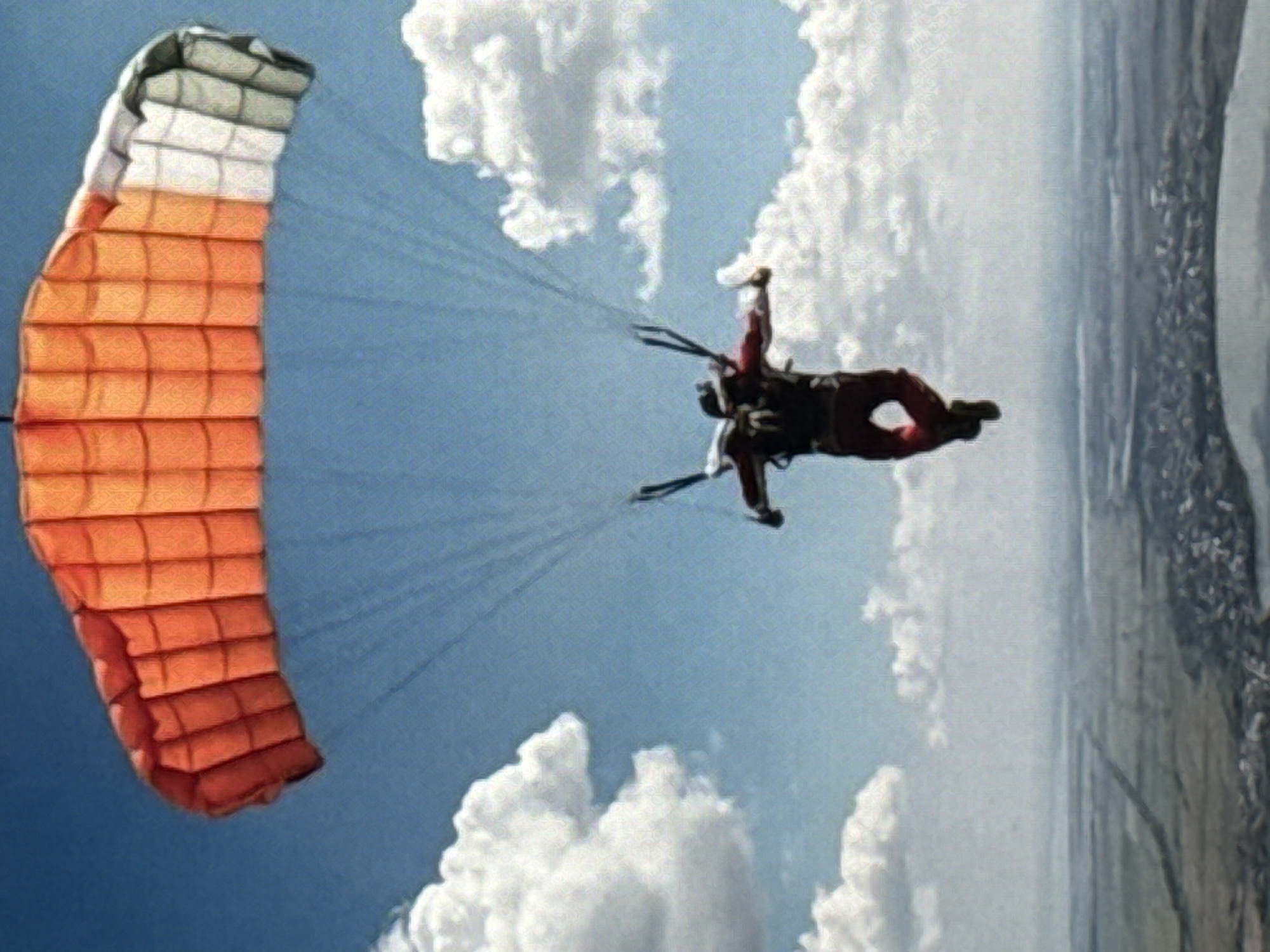 A skydiver is captured mid-air, descending under a multi-colored parachute against a bright blue sky dotted with white clouds. The parachute is predominantly orange with white and grey sections. The skydiver, wearing a red and black suit and a helmet, is positioned in the center of the frame, with legs extended downwards. Below the skydiver, a vast, hazy landscape unfolds, featuring a city with buildings, roads, and what appears to be a body of water or a large plain. The overall impression is one of extreme sport and aerial activity above a sprawling urban and natural environment.