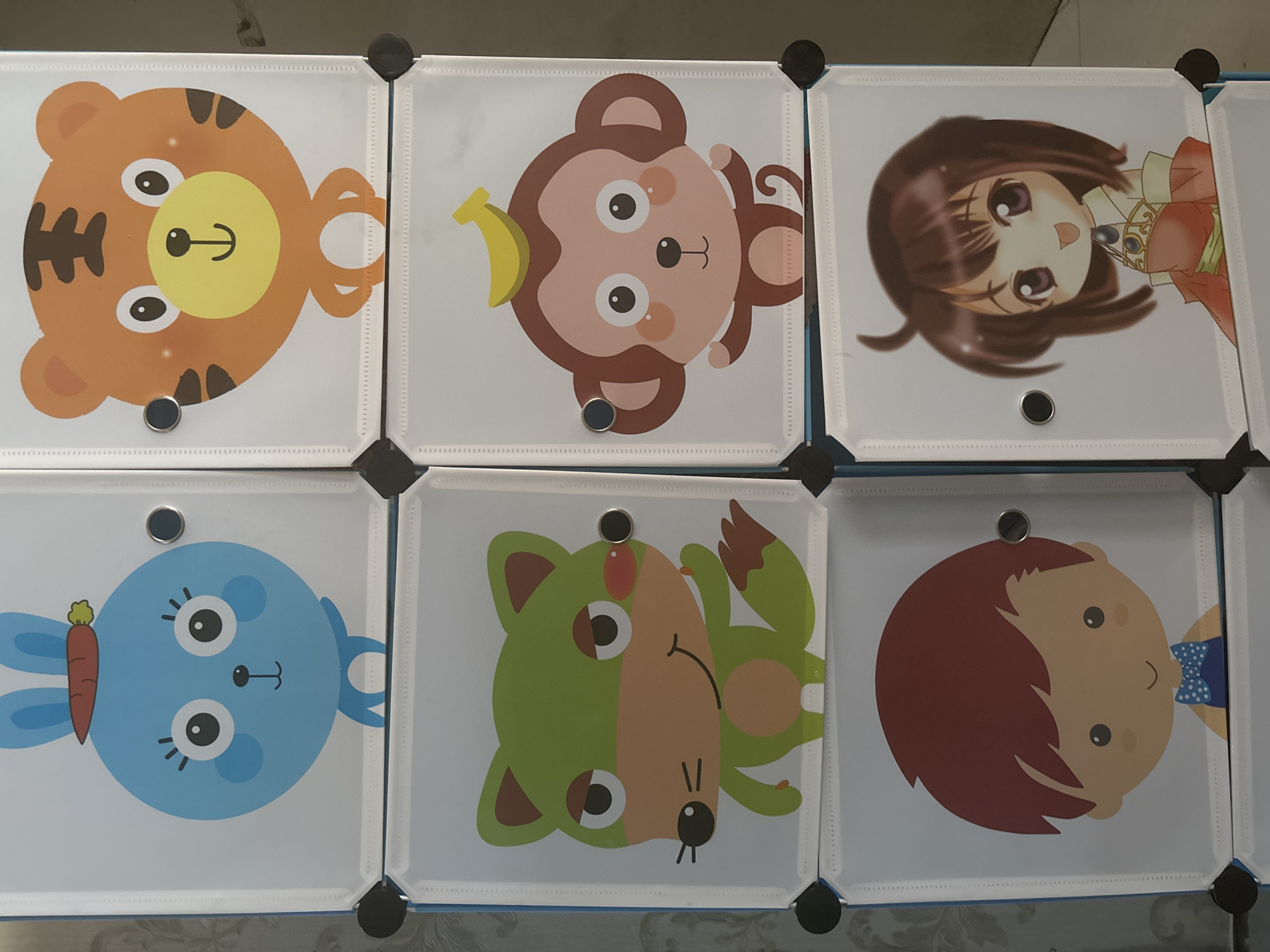 The image shows a modular storage unit with six doors, each decorated with a cartoon character. The unit is positioned against a textured, light-colored wall. From top-left, clockwise, the characters are: a blue bunny with a carrot on its head, an orange tiger, a monkey with a banana on its head, a chibi-style anime girl in a red dress, a boy with brown hair and a blue bowtie, and a green fox. The storage unit is likely located in a home or a child-oriented space, such as a playroom or bedroom. The context does not provide any specific location within Tangerang, Indonesia.