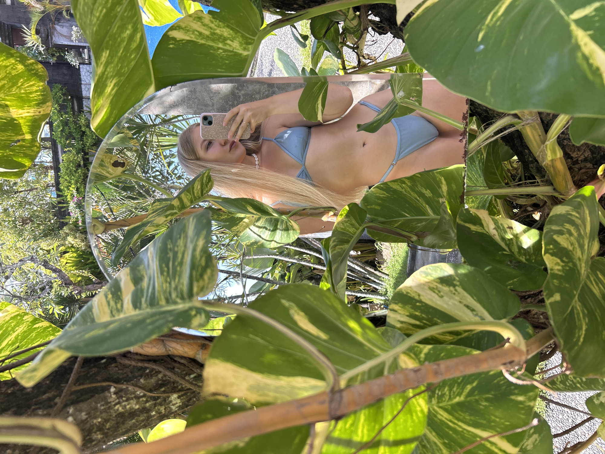 A mirror selfie shows a woman in a light blue bikini, surrounded by green and yellow variegated leaves, potentially Golden Pothos, affixed to a tree or structure. The mirror is positioned amidst foliage, providing a view of the surroundings. In the background, there is a partial view of trees, a building with a lattice structure, and the sky. Based on information provided, this scene may be located in Kuta Selatan, Indonesia.