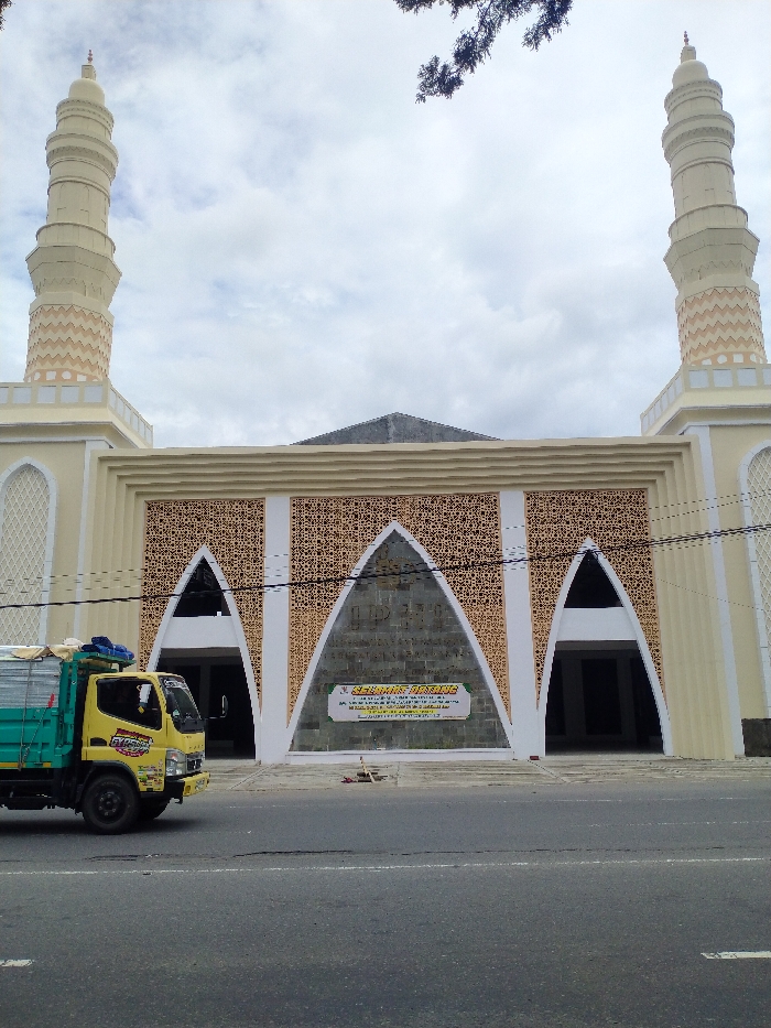 A daytime exterior shot captures a mosque and a truck in Karanganyar, Indonesia, under an overcast sky. The prominent subject is a large, light-colored mosque featuring two tall, slender minarets flanking its facade. The mosque's front is adorned with intricate geometric patterned panels, predominantly above three large arched entrances. The central arch is the widest and set back, containing a banner. The two side arches lead into shaded areas.
In the foreground, a yellow delivery truck with a green open bed is seen moving or parked on an asphalt road. The truck's cab has "BYPASS" written on its door. Overhanging branches of a tree are visible in the upper left corner, and power lines cross horizontally across the middle of the frame.
The banner within the central archway of the mosque prominently displays "SELAMAT DATANG" (Welcome) in large letters, with smaller, less legible text underneath, likely providing further information or a message. The mosque appears to be a new or recently renovated structure. The scene depicts a typical street view in an urban or semi-urban setting.