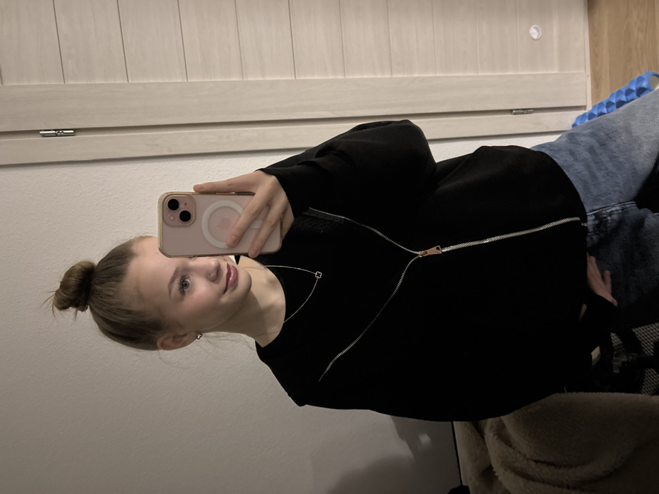 A young woman takes a selfie. She has blonde hair pulled into a bun, blue eyes, and a silver nose piercing. She wears a black zippered sweatshirt and jeans. A clover-shaped pendant hangs on a delicate silver necklace. Her iPhone has a light pink case. A door with horizontal panels is behind her, and what appears to be a blue massage roller is on the floor nearby. There are also some indistinct objects in the foreground.