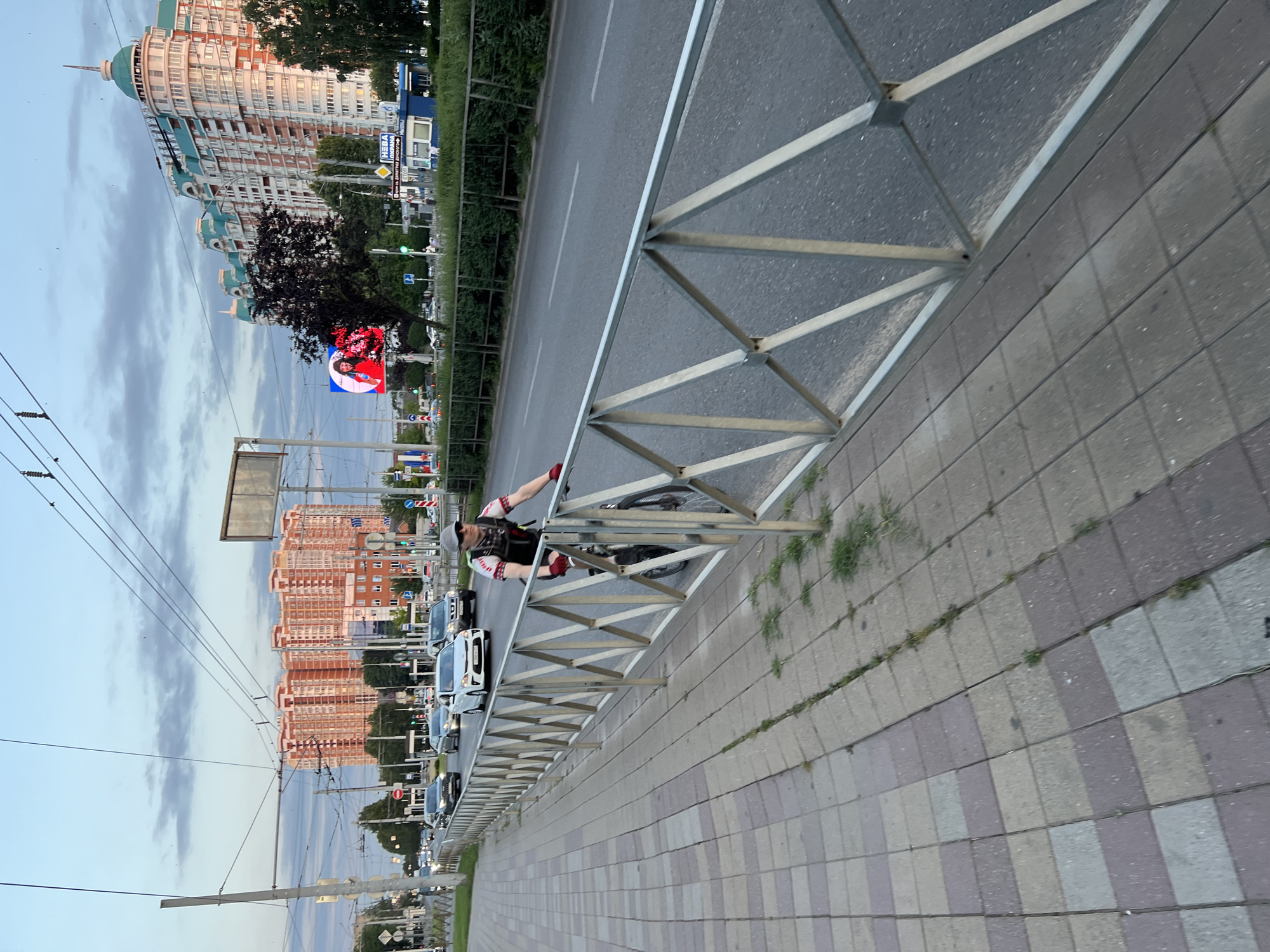 The image captures a street scene in Krasnodar, Russia, at what appears to be dusk. The perspective is from a sidewalk, looking across a metal fence towards a road with moderate traffic. A person is cycling on the sidewalk, wearing a backpack and cycling gear. The road is lined with trees and features several cars in motion. In the background, prominent residential buildings with distinctive architecture dominate the skyline. One building is tall with light-colored facades and teal-colored roofs and spires, while another complex is a large, multi-story, pink-hued building. A digital billboard displaying a woman is visible on the left side of the road. Overhead, power lines crisscross the sky, indicating an urban environment with a public transportation system. The sidewalk is paved with square tiles, and weeds are growing through the cracks. The sky is a soft blue with hints of clouds, typical of twilight.