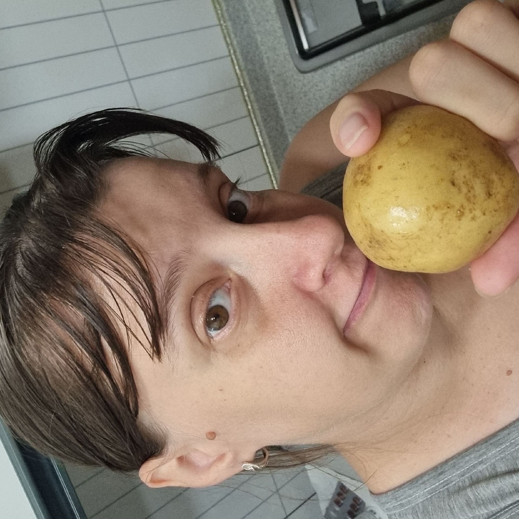A woman is holding a potato in front of her face, with her left eye visible and her mouth partially obscured by the potato. She is wearing a grey t-shirt and a silver hoop earring. Her hair is dark brown and pulled back. The background shows a tiled wall and a grey countertop, suggesting a kitchen environment. The lighting is natural and appears to be coming from the front. The image was taken indoors.