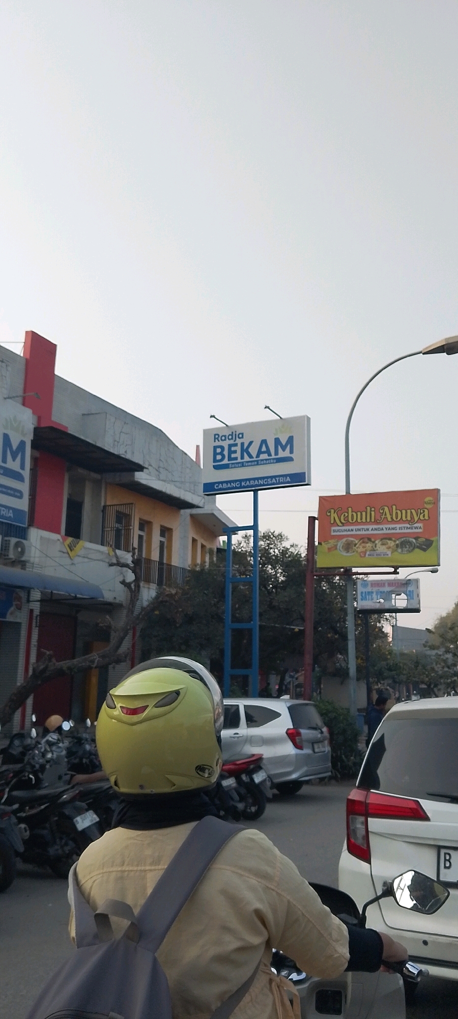 In Karangsatria, Indonesia, an outdoor daytime scene features a person wearing a yellow helmet and a beige jacket on a motorcycle, viewed from behind. They are on a street with several parked motorcycles to their left and various vehicles, including a silver car and a white car, on the right. In the background, buildings and billboards are visible. One prominent billboard displays "Radja BEKAM Solusi Teman Sehatku CABANG KARANGSATRIA" in blue and white text. Another billboard, orange with yellow accents, advertises "Kebuli Abuya" with images of food. A smaller sign below it reads "RUMAH MAKAN SATE BEBEK" and "SATE BEBEK JOGJA". The environment suggests a commercial area with a clear sky overhead.