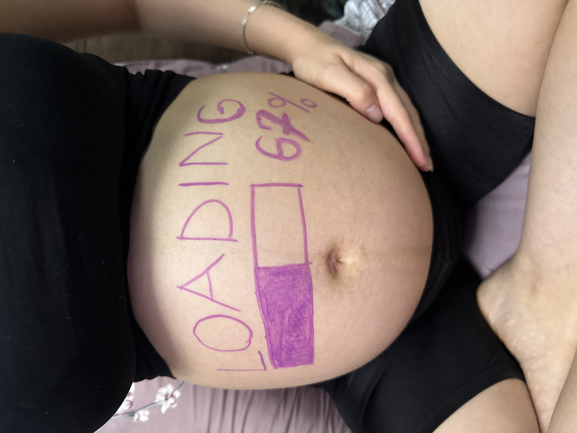 The image shows a close-up of a pregnant person's belly, with "LOADING 67%" written on it in purple marker. A progress bar, also drawn in purple marker, is shown partially filled. The person is wearing black clothing. The setting appears to be indoors, with a hint of a floral patterned bedspread. The image is likely a snapshot taken by the pregnant person themselves, intended to be shared as a pregnancy update or announcement. The mood is playful and anticipatory. The text visible in the image is "LOADING 67%".