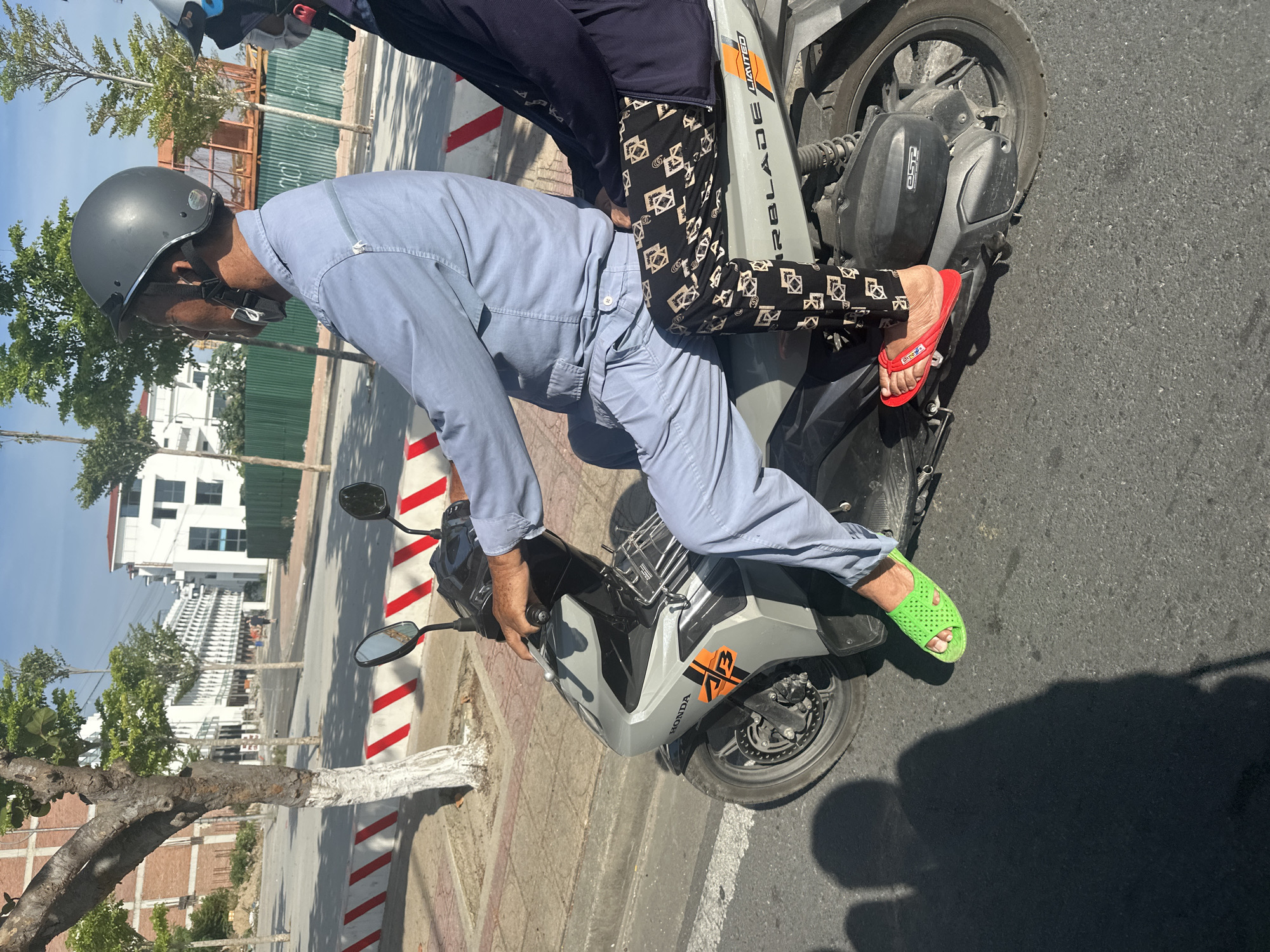 In Nha Trang Ward, Vietnam, two individuals are riding a Honda Airblade scooter on an asphalt road. The driver, wearing a light blue long-sleeved shirt, light blue pants, a dark gray helmet, and green rubber slides, is operating the scooter. The passenger behind the driver, wearing a dark navy long-sleeved top and black pants with a geometric pattern, has their feet resting on the scooter. The scooter is gray and black with "HONDA" and "AIRBLADE LIMITED" branding visible. In the background, there are trees, buildings under construction with scaffolding, and a green fence. The road has a white and red striped curb. The lighting suggests it is daytime, with distinct shadows cast on the road.
