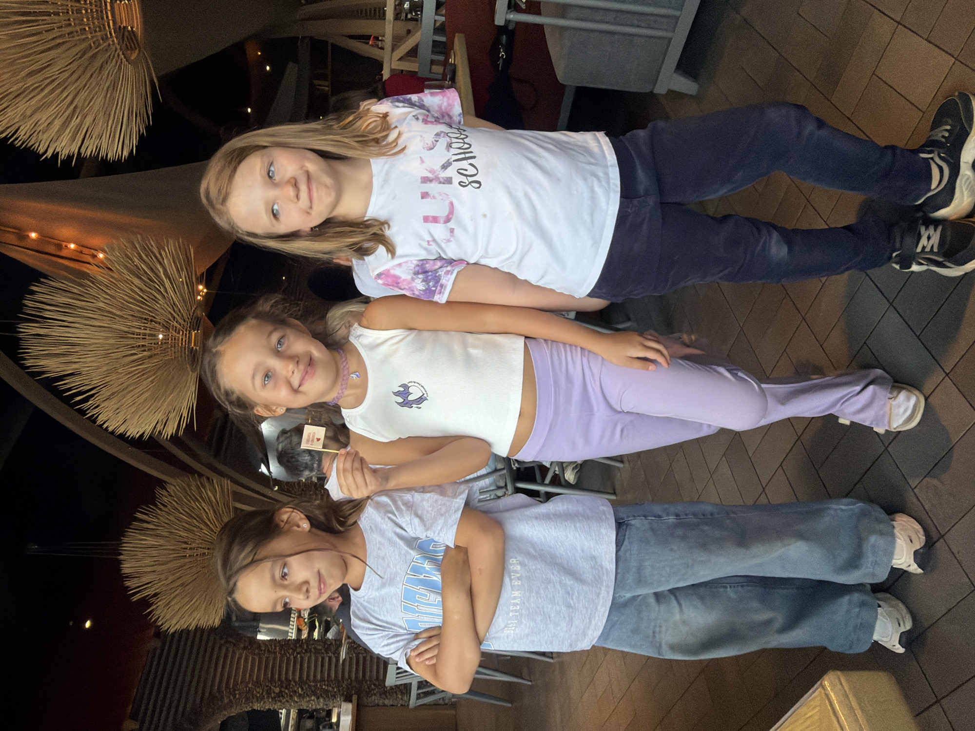 Three young girls stand indoors beneath what appears to be decorative, spiky light fixtures. The eldest girl wears a white t-shirt with "LKS School" printed on it, paired with dark blue pants and sneakers. The middle girl wears a white top, light purple pants, and sneakers. She holds a small flag in her hand. The youngest girl wears a light blue t-shirt and blue jeans, also with sneakers, and has her arms crossed. They are likely standing in a restaurant or dining area, considering the presence of chairs and tables visible in the background. The location in Krasnoyarsk, Russia is not possible to confirm from the image alone.