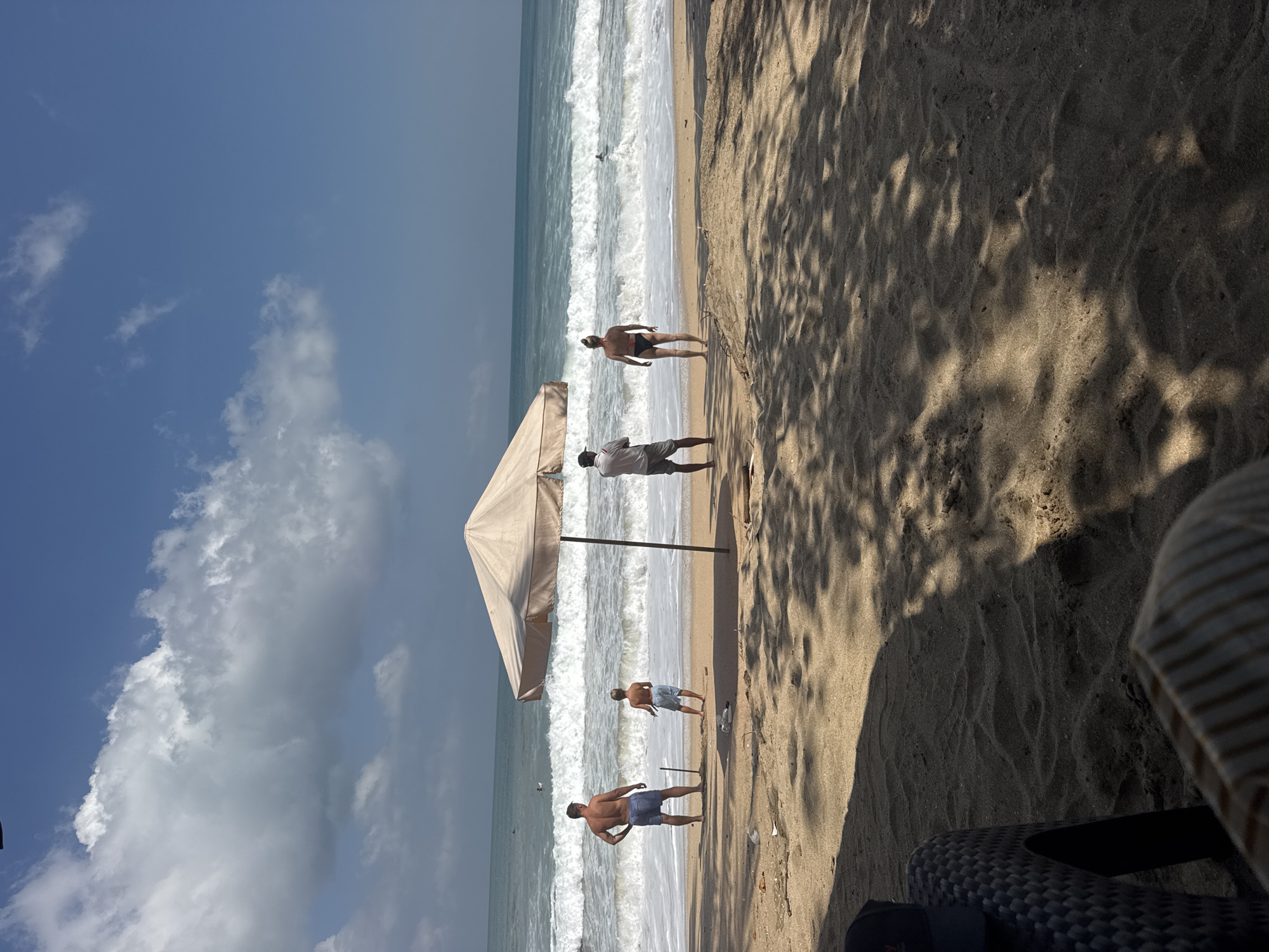 The image depicts a beach scene in Kuta, Indonesia, likely during the daytime given the bright sunlight casting shadows on the sand. The ocean is active with moderate waves crashing onto the shore, suggesting a lively atmosphere for water activities. Several people are visible: two men in swim trunks are standing near the water's edge, one with his hands on his hips and the other looking out at the sea. A third man, wearing shorts and a white t-shirt, stands near a large, light-colored beach umbrella, possibly attending to beachgoers or working. Further out in the water, another person can be seen swimming or wading.
The setting is a sandy beach with a clear blue sky dotted with white clouds. The shadows of palm trees or foliage create a dappled pattern on the sand in the foreground. Beach chairs, one partially visible and appearing to be made of woven material, suggest a resort or public beach area. The overall mood appears relaxed and recreational, consistent with a beach vacation or day at the shore. There is no visible text in the image.