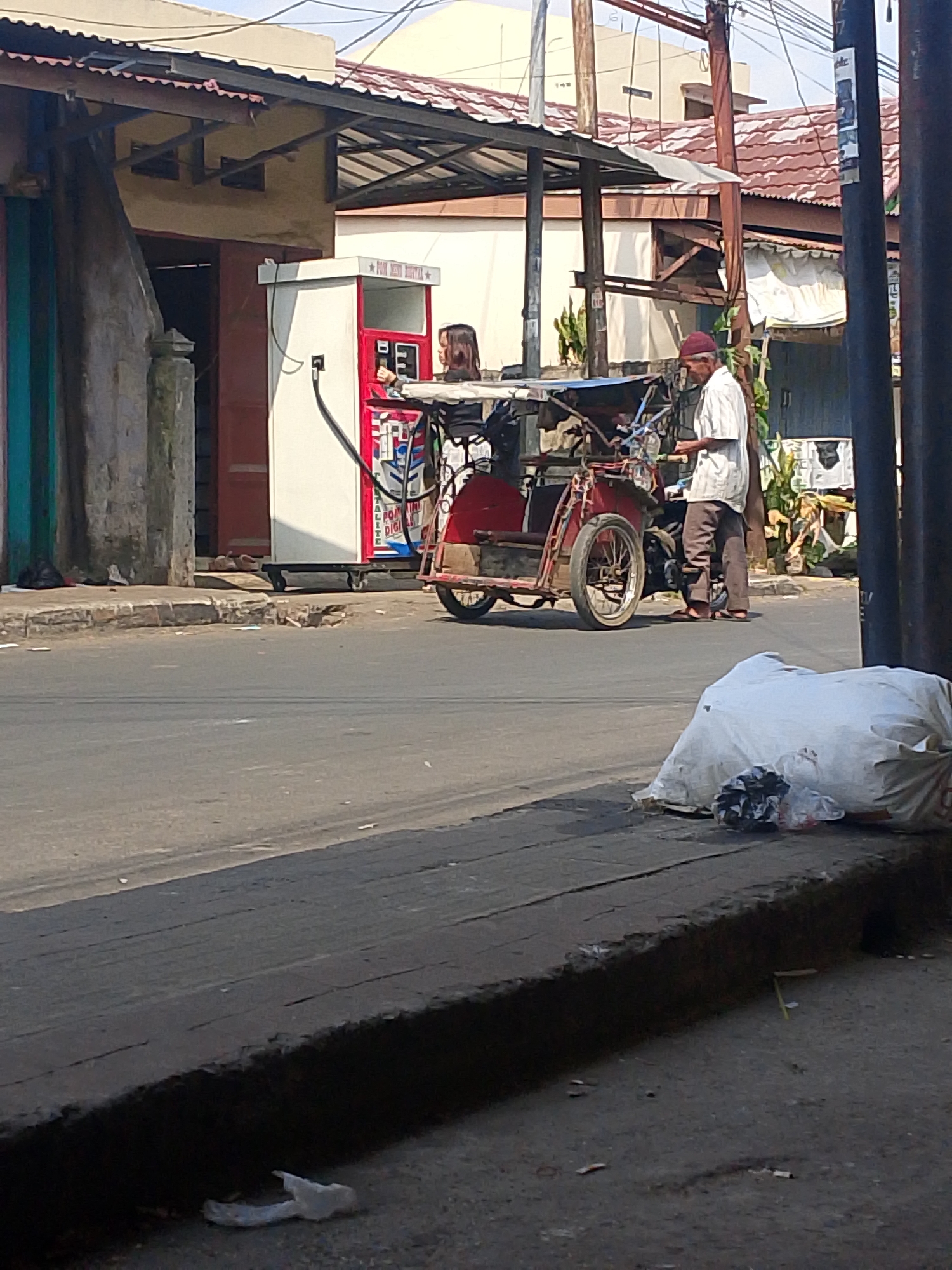 This is a medium-shot, eye-level, outdoor photograph of a street scene in Palembang, Indonesia, under daylight conditions. The foreground features a raised concrete curb with dark, rough-textured planks or asphalt laid on top, bordering a grey asphalt road. A large white trash bag filled with what appears to be plastic and other refuse is partially visible on the curb. Scattered debris, including paper and what looks like cigarette butts, is present on the road and curb.
In the mid-ground, a red and white gas pump stands to the left, with a hose connected to a mobile red and silver three-wheeled vehicle, likely a modified motorcycle or auto-rickshaw. A young woman with dark hair, wearing a black top and patterned pants, stands by the gas pump, interacting with it. Further to the right, an older man with a red head covering and a light-colored, short-sleeved shirt and brown pants stands next to the three-wheeled vehicle, holding something in his hand. The vehicle itself has a covered seating area and exposed mechanical components.
The background shows buildings with red-tiled roofs and awnings supported by poles. A tall, dark pole with wires running from it is positioned on the right side of the frame, obscuring a portion of the background. The overall environment suggests a commercial or residential street in a town or city. The lighting indicates a sunny day with distinct shadows.