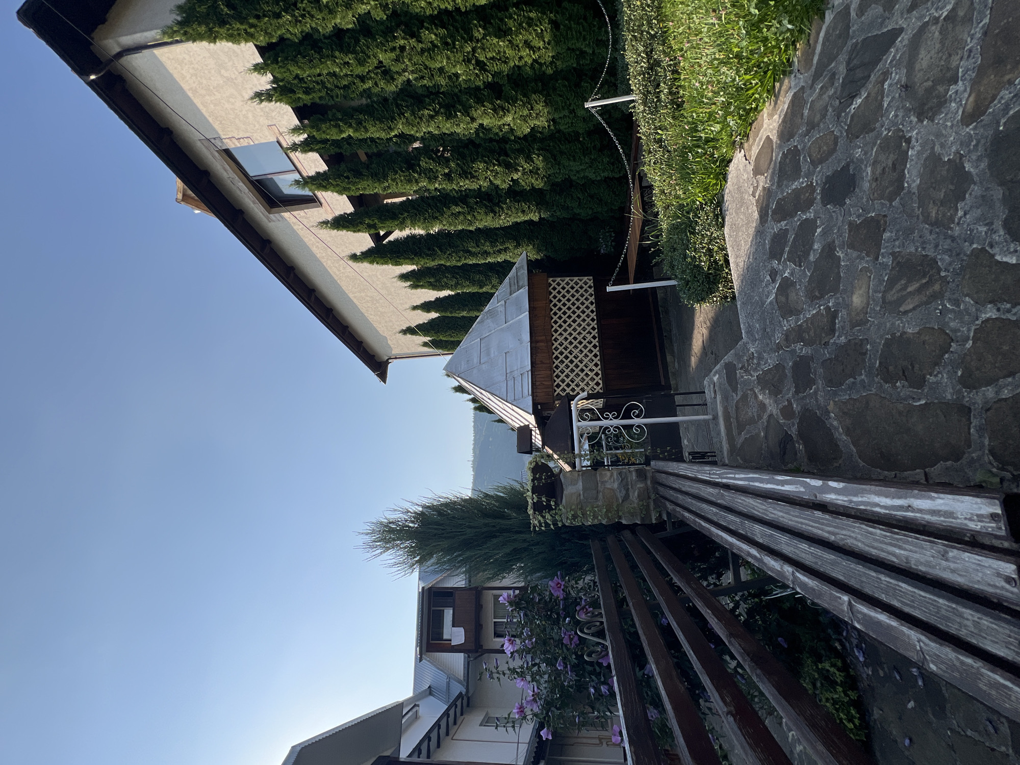 The image depicts a residential area in Yaremche, Ukraine, under a clear blue sky. In the foreground, a rustic wooden bench is positioned on a stone pathway. To the left, a wooden fence borders a garden with flowering bushes. Beyond the fence, a portion of a white building with a sloped roof is visible, and next to it, a taller, multi-story building features a balcony with a towel hanging over the railing. A tall, slender evergreen tree stands prominently near the center of the frame. To the right, a small, ornate gazebo with a hexagonal roof and latticework sides is situated. A row of tall, dark green evergreen trees lines the side of a light-colored, textured building that occupies the right side of the image. Green bushes and ground cover are visible in the middle ground and foreground right, with a stone pillar supporting a potted plant. In the background, a distant hillside is visible. The overall scene suggests a peaceful, well-maintained outdoor space, likely a courtyard or garden area of a private residence or guest house.