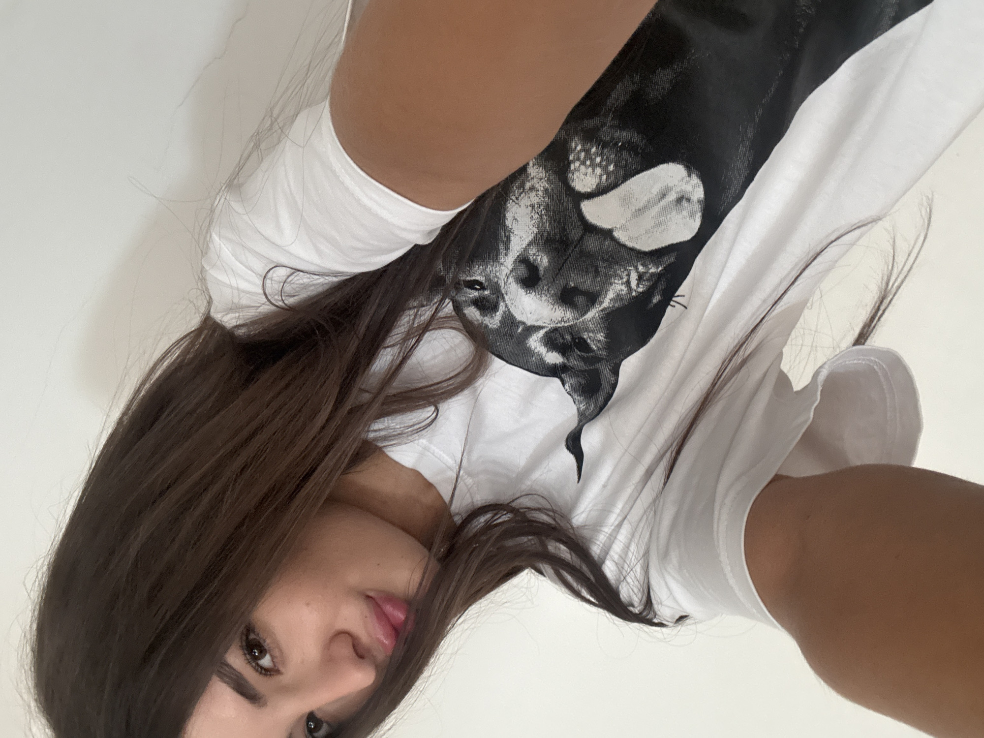 A young woman with long, dark hair is taking a selfie. She is wearing a white t-shirt with a black and white graphic of a dog's face. The background is a plain white wall. Her face is close to the camera, and she is looking slightly to her left with a slight smile and her lips slightly parted.
