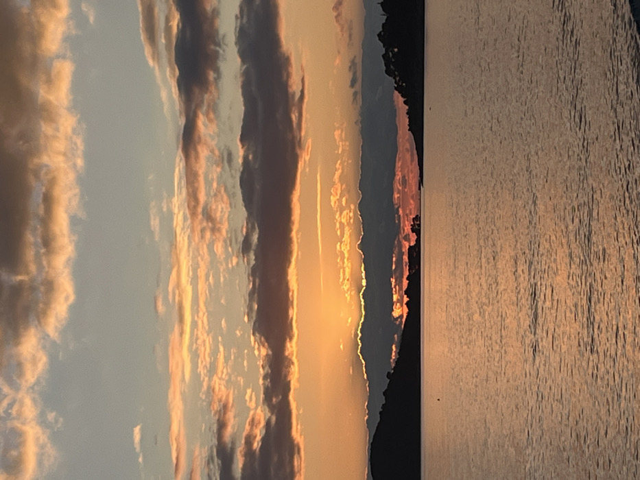 The scene depicts a coastal sunset observed from Rafailovići, Montenegro. The sky occupies the upper half of the image, displaying a gradient from light blue at the zenith to vibrant orange and yellow directly above the horizon. Multiple cloud formations are present: scattered, elongated altocumulus clouds tinted with orange and pink are visible across the upper and middle sky, while a dense, elongated band of dark grey stratocumulus clouds stretches horizontally across the mid-sky. The setting sun, a bright orange disc, is partially obscured behind this dark cloud band, emitting a strong glow.
Below the sky, silhouetted landmasses, appearing as dark, undulating hills or coastline, frame the central view. These landforms are visible on both the left and right sides, with discernible tree lines atop them. The lower half of the image consists of a calm body of water, likely the Adriatic Sea or a bay, reflecting the warm hues of the sunset. The water's surface exhibits subtle ripples, with colors ranging from deep orange and brown near the horizon to darker tones in the foreground. A single, dark, cylindrical buoy is present in the water, located approximately one-third of the way from the left edge of the frame. No individuals, vessels other than the buoy, or explicit actions are observed within the frame.