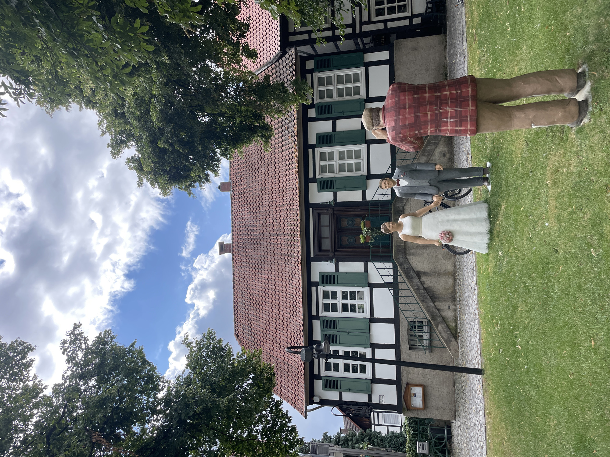 In Salzkotten, Germany, in front of a traditional half-timbered house with a red-tiled roof, two life-size figures of a bride and groom stand on a patch of grass. The bride wears a white wedding dress and holds a bouquet of pink flowers, while the groom is dressed in a gray suit with a red bow tie. To the right, a third life-size figure, dressed in a red plaid shirt and brown pants, stands with its back to the camera, appearing to take a photograph of the couple. The house features white walls with dark timber framing, green shutters on the windows, and a prominent entrance with a staircase leading up to it. Lush green trees frame the scene, and the sky above is partly cloudy with blue patches.