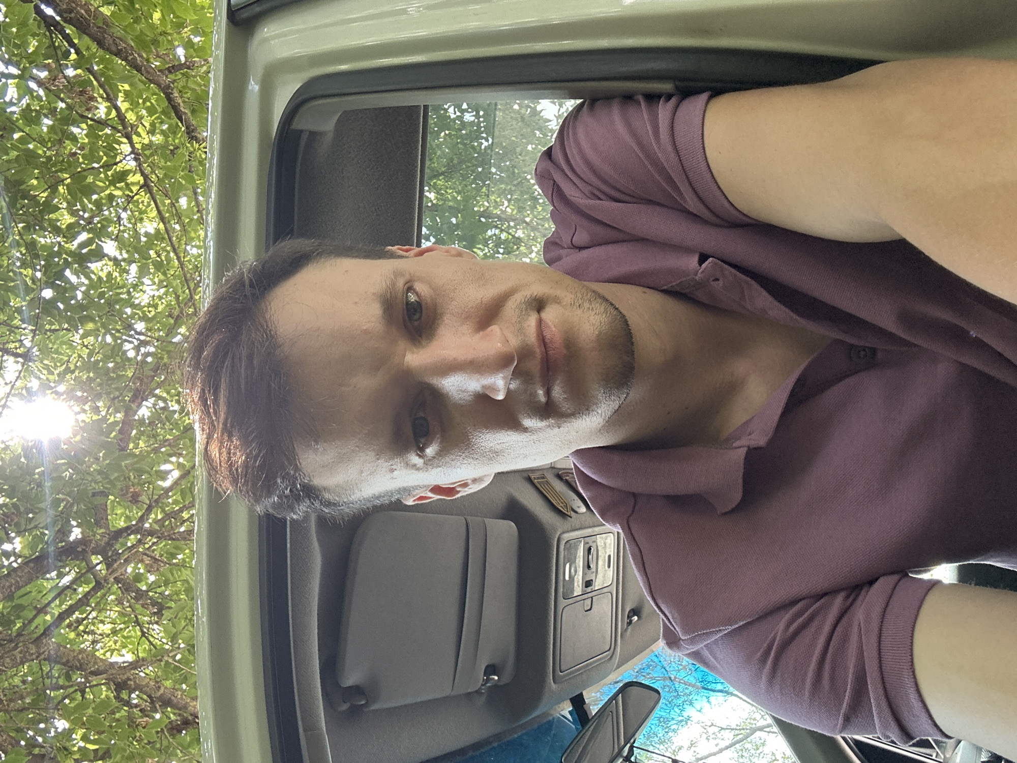 The image shows a man in his late 20s or early 30s, with short brown hair and light brown eyes, looking directly at the camera. He is wearing a short-sleeved, collared, plum-colored polo shirt. The man is positioned inside a vehicle, with the interior of the car visible, including the sun visor and roof console. The exterior of the vehicle, and the outside environment, are visible through the car window, with trees and sunlight suggesting an outdoor setting. The image appears to be a selfie taken from within the car. There is no information in the image to determine the specific location within Makiv, Ukraine.