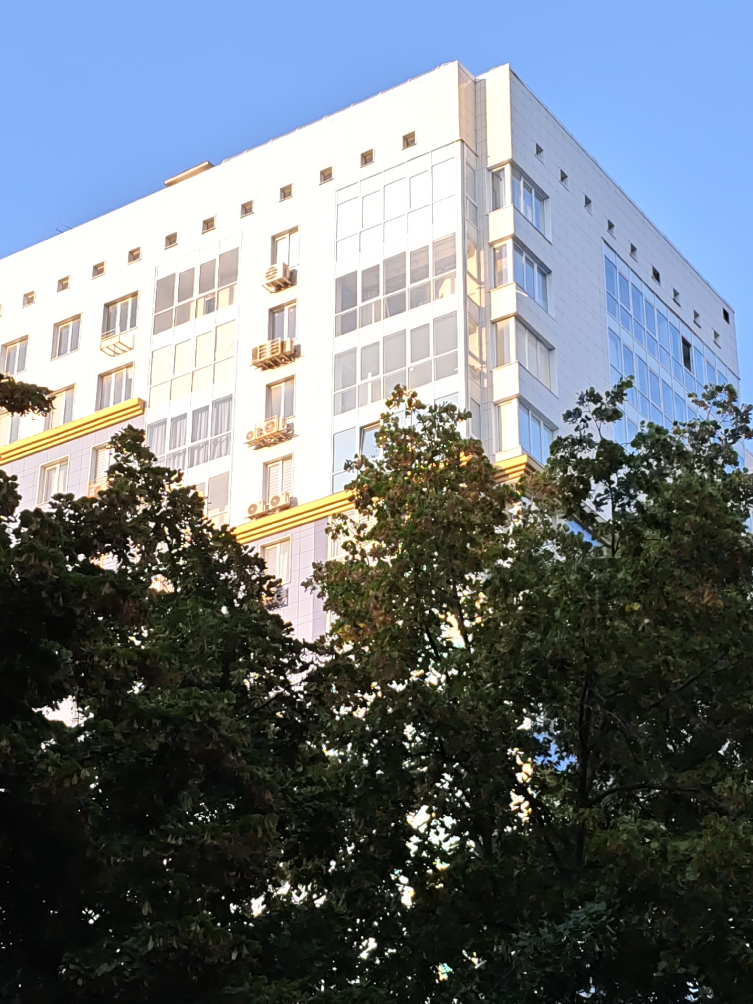 This media file features a modern, multi-story white building against a clear blue sky. The building has numerous windows, some of which are large and reflective, indicating a bright, sunny day. Several air conditioning units are visible mounted on the exterior walls.
In the foreground, lush green trees with full foliage partially obscure the lower floors of the building. The leaves of the trees are dark green, suggesting a healthy canopy. The lighting suggests it is daytime, likely late afternoon or early morning, given the warm sunlight casting a yellowish glow on parts of the building and through the trees. The scene depicts an urban or suburban setting, with the building serving as a prominent architectural element. There is no visible text in the image. The overall impression is one of a calm, everyday scene in a well-maintained environment.