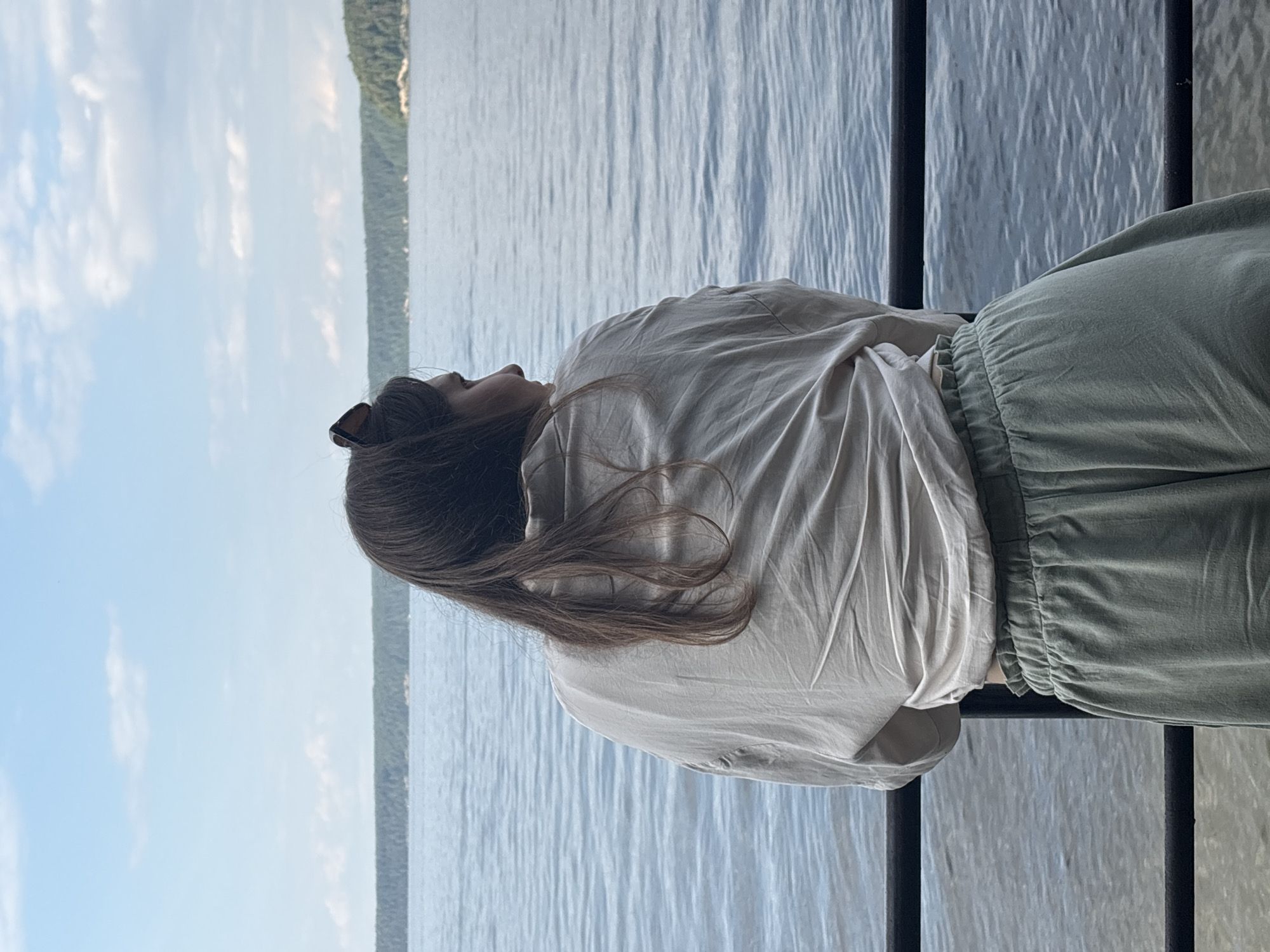 A woman stands on a walkway, her back to the camera, facing a wide body of water. She has long, dark brown hair and is wearing a loose, light-colored shirt and pale green pants. Her head is turned to the right, and she appears to be looking out at the water. Sunglasses are perched on top of her head. The water stretches out to a tree-lined horizon, with a pale blue sky with scattered clouds above. The location is the shore of a large lake or river in Lyady, Russia.
