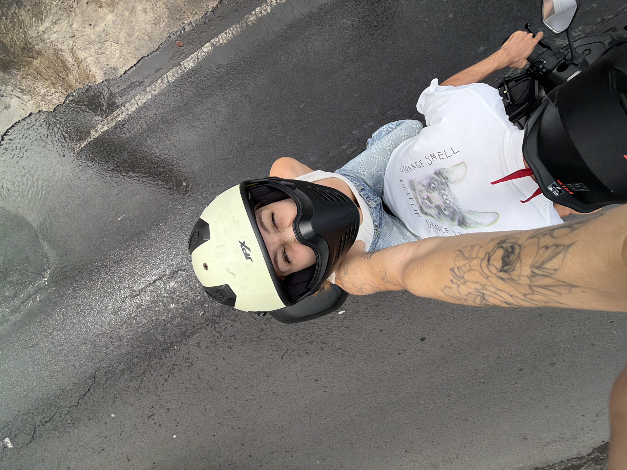 A first-person perspective shot captures two individuals on a motorcycle, likely on a wet road in North Kuta, Indonesia. The person in the foreground, whose arm is visible with a tattoo of a woman's face and flowers, is operating the motorcycle. In the background, a woman wearing a light-colored helmet with "JPX" branding and a black face shield, a white tank top, and light blue jeans is seated behind the driver. She is looking up at the camera with a playful expression. The motorcycle has a black helmet on the back, adorned with a red tag and a sticker that reads "MX 722-R DOT". The road surface is asphalt, with visible cracks and a thin white line marking the lane. The surrounding environment appears to be outdoors, with the road showing signs of recent rain, indicated by the wet sheen.