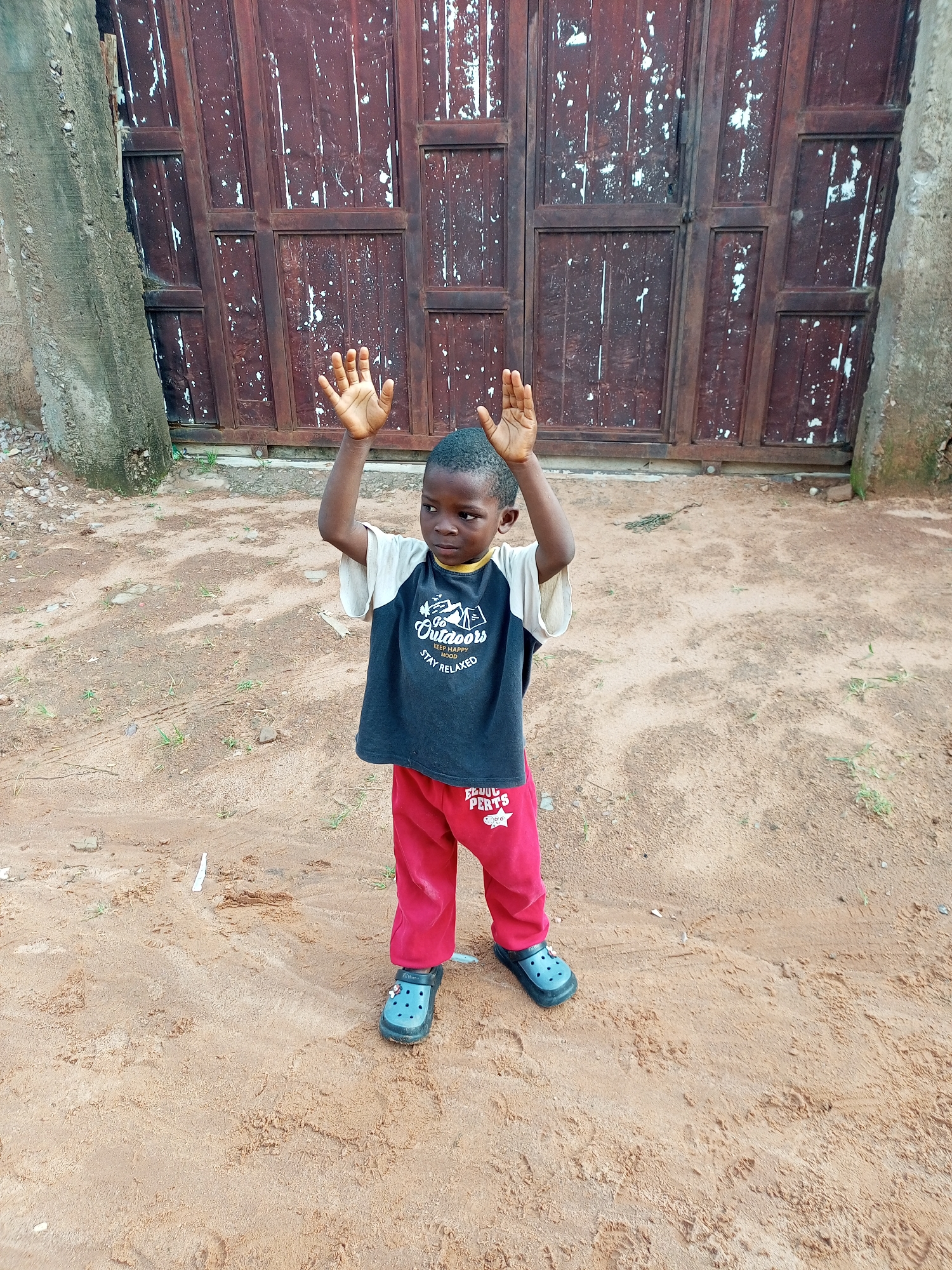 A young Black boy stands in a sandy, outdoor area in front of a large, weathered wooden gate. The boy is wearing a dark t-shirt with a graphic that includes the words "Go Outdoors," "Keep Happy Mood," and "Stay Relaxed." He also wears red pants and blue clogs. His hands are raised, and he appears to be looking off to his right. The gate behind him is dark brown with lighter splotches, suggesting age and wear. The ground is sandy with some sparse green vegetation. The overall impression is a simple, everyday scene.
