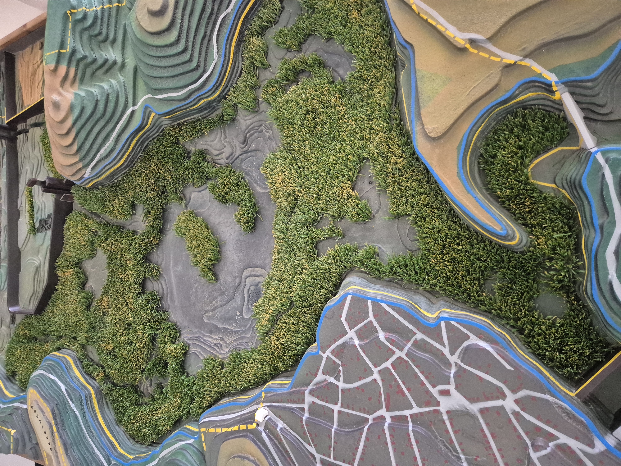 The image depicts a topographic model of the Srebarna area in Bulgaria. The model uses layered contours to represent elevation, with green artificial turf indicating forested or vegetated areas. Blue and yellow lines likely represent bodies of water or roads. One section of the model features a grey surface with a white grid pattern and red splotches, potentially indicating a built-up area or urban zone within the Srebarna region. The model is built into a frame, and is likely displayed as a educational or informational exhibit.