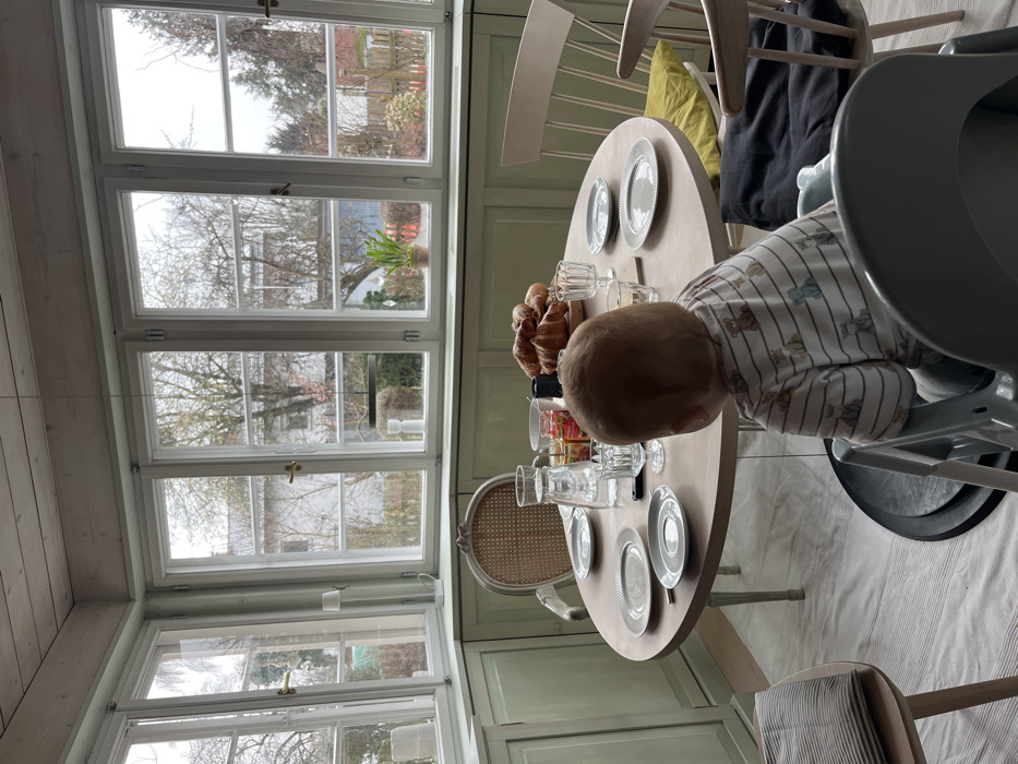 The image displays an indoor dining area or sunroom in Hohenbrunn, Germany. A round, light-colored wooden table is centrally positioned, set with five light-grey plates, a clear glass pitcher of water, multiple clear drinking glasses, small jars of spreads, a dark cylindrical object, and a pile of croissants on a wooden board. An infant, wearing a striped long-sleeved shirt, is seated in a light-grey high chair, with its back to the camera, facing the table. Two additional wooden chairs are visible around the table; one has a cane back, and the other a slatted back with a yellow cushion. Another wooden chair with a striped cushion is in the left foreground.
The room features numerous large, white-framed windows that form a corner, providing ample natural light. The walls below the windows are paneled and painted a light mint green, contrasting with the light-colored wooden plank ceiling. Outside the windows, a residential neighborhood is visible, characterized by bare deciduous trees, some evergreen foliage, and distant house structures under an overcast sky. A small potted plant rests on the central windowsill, and a white floor lamp is visible in the left corner near the window. A black suspended light fixture hangs from the ceiling, visible through the windows from the inside. The floor is covered by a light-colored rug or painted surface with subtle striped patterns.