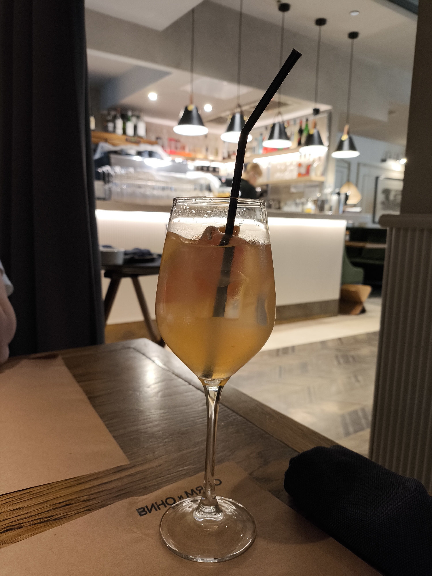 This image presents an indoor scene from the "Вино и Мясо" restaurant, located at Komunnorov, 218, Izhevsk, Russia. The foreground is dominated by a clear, stemmed wine glass holding a light orange-yellow beverage, indicative of a cocktail like an Aperol Spritz, with several ice cubes and a black bent plastic straw inserted. This glass rests on a dark wooden table, which features a prominent grain. Beneath the glass and extending across the table is a light brown paper placemat, with partial Cyrillic text "ВИНО И МЯ" (meaning "Wine and Mea," likely "Вино и Мясо" for "Wine and Meat") printed in dark lettering. To the immediate right of the glass, a dark, tightly rolled fabric napkin is positioned on the placemat.
The background is softly blurred but reveals distinct elements of a contemporary restaurant or bar. To the left, a dark curtain or drapery is partially visible. The central background features a modern bar area characterized by a luminous white counter. Behind this counter, shelves display an array of liquor bottles, various glasses, and other bar equipment. Overhead, multiple black, conical pendant lights illuminate the bar area. An indistinct human figure, appearing to be a staff member, is faintly visible behind the bar. To the far right of the frame, a vertical architectural element, possibly a column or wall panel with fluted detailing, is present. The floor, visible beyond the table, features a light-colored, possibly tiled, surface with a geometric pattern. The ambient lighting is dim but functional, consistent with an evening dining establishment. No specific actions or direct interactions are occurring within the visible frame.