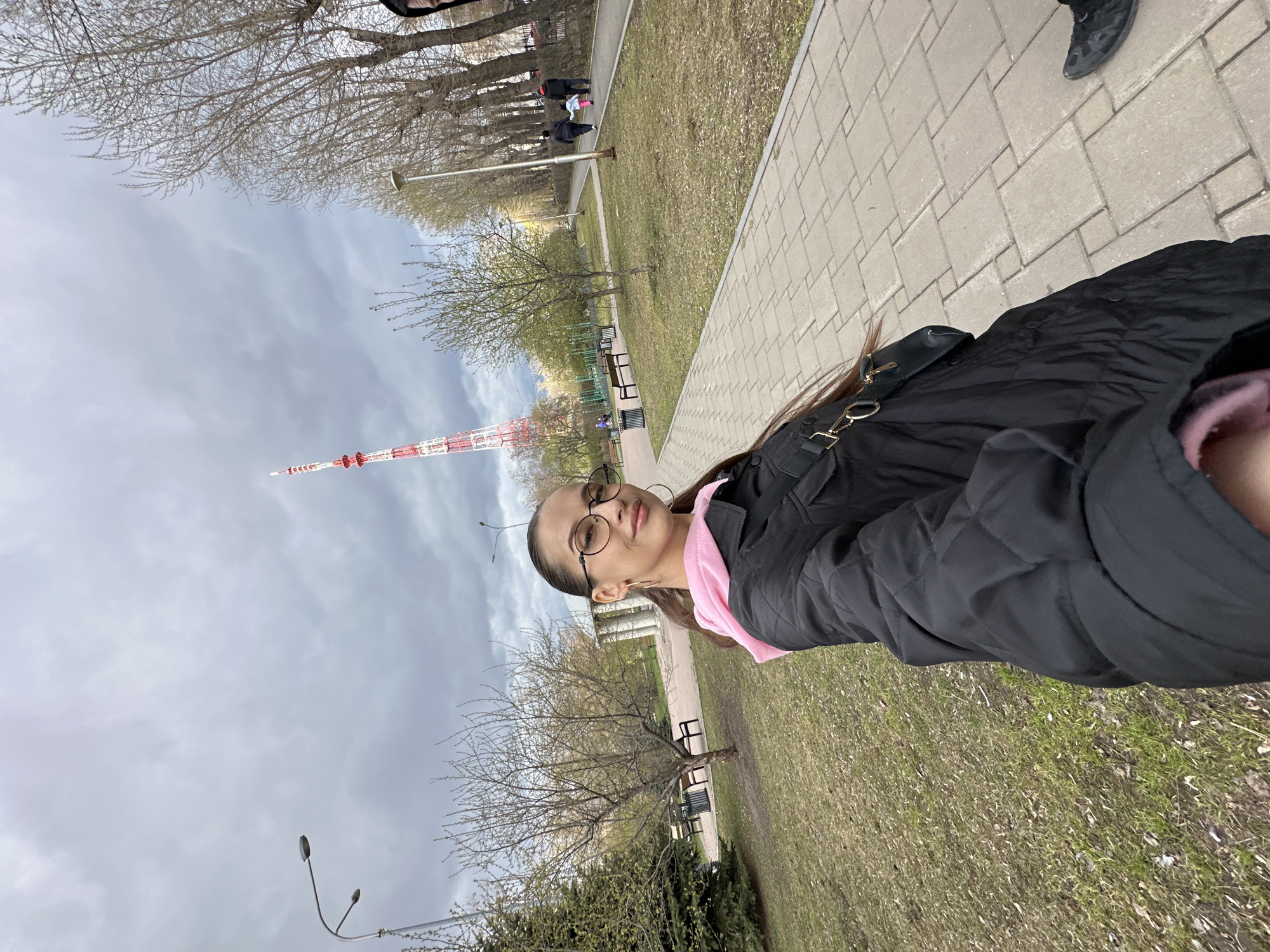 A person is posing for a selfie along a paved walkway in a park in Yekaterinburg, Russia. The subject wears glasses, a pink garment, and a black jacket. A black purse hangs on the subject's shoulder. In the background, trees line the walkway, and a television tower is visible. The sky is overcast. Other people are walking in the background. There are park benches on the grass near the walkway.
