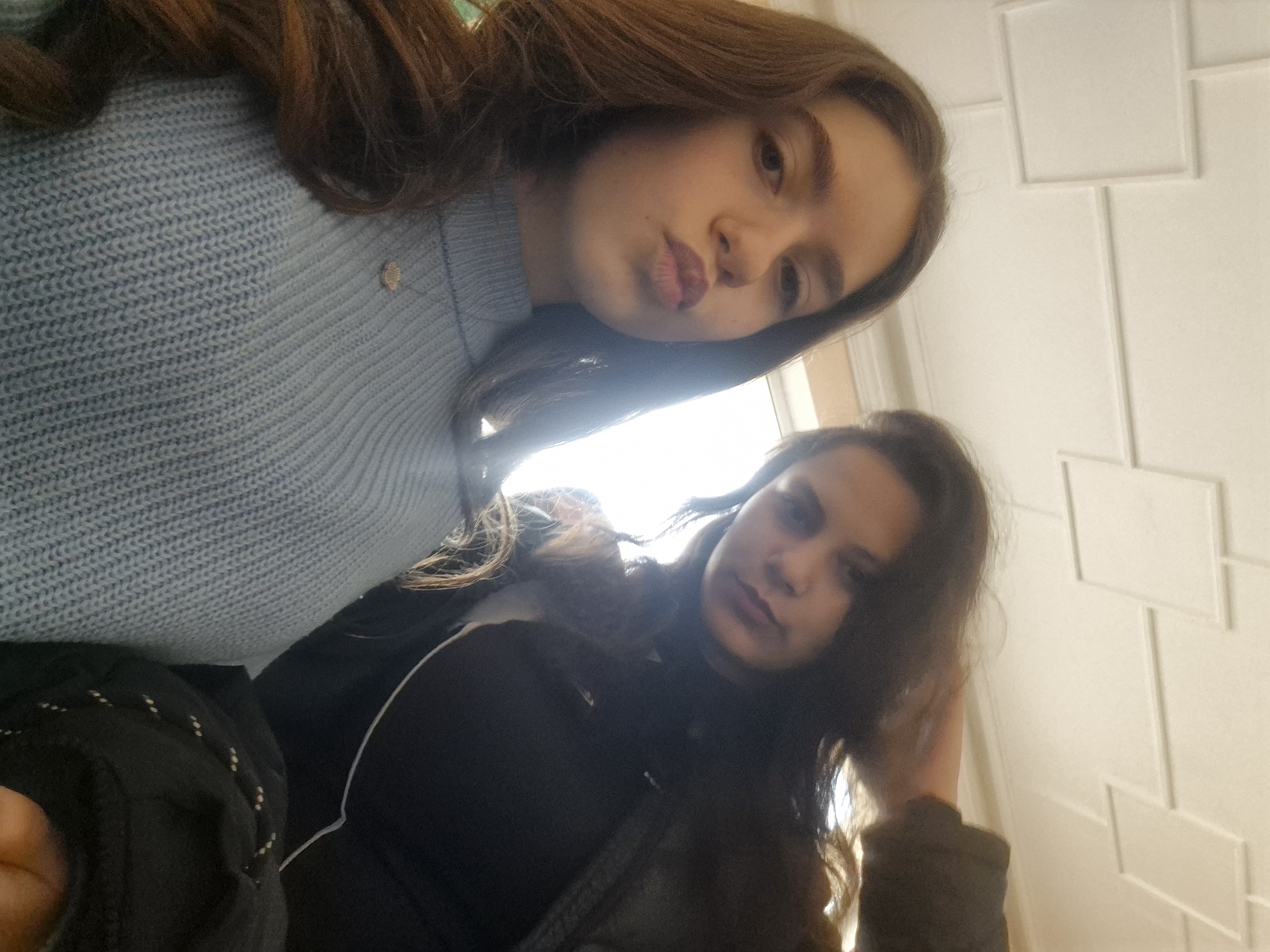 Two young women are in a selfie. The woman on the left is wearing a light blue sweater and has brown hair. The woman on the right is wearing a black top and has brown hair. The background is a white wall with a geometric pattern. The location is unknown and cannot be determined.