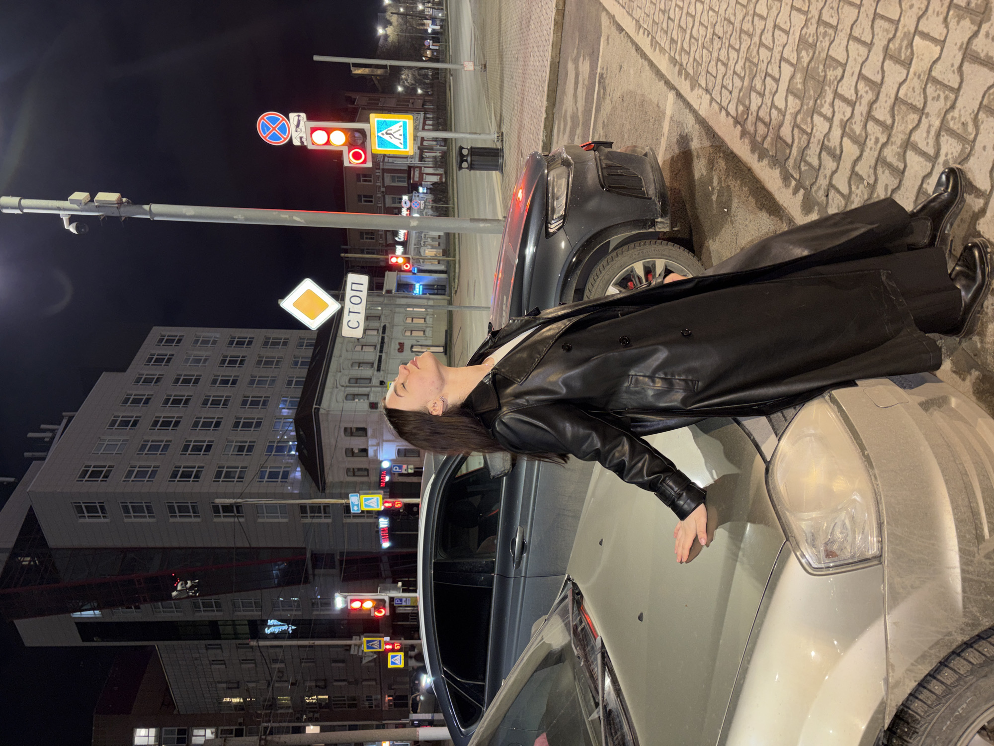 A young woman in a black leather coat and boots is leaning on the hood of a silver car on a city street at night. In the background are buildings, traffic lights, and street signs. The signs are in Russian, one reading "СТОП," which translates to "Stop." A pedestrian crossing sign is also visible. Based on the architecture and street signage, the location is the city of Perm, Russia. Another dark-colored car is parked nearby. The pavement is brick.