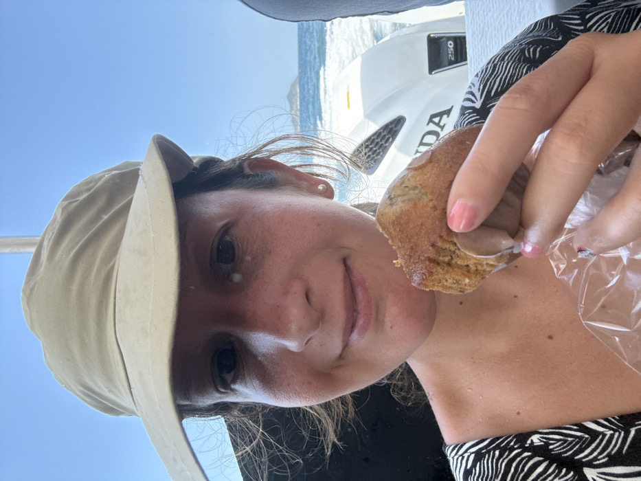 A female individual is depicted in a close-up, chest-up shot aboard a vessel. She wears a beige wide-brimmed sun hat and a black and white patterned top. Her face shows a slight smile, with a small white smudge present below her right eye. She holds a partially consumed, light brown muffin, wrapped at its base in clear plastic, in her left hand, which exhibits pink-painted fingernails. In the midground to her right, a white Honda 250 horsepower outboard motor is visible. The background reveals choppy blue water leading to a distant, hazy landmass under a clear, bright blue sky. The scene is consistent with a coastal or marine environment, specifically in the city of Pak Nam, Thailand.