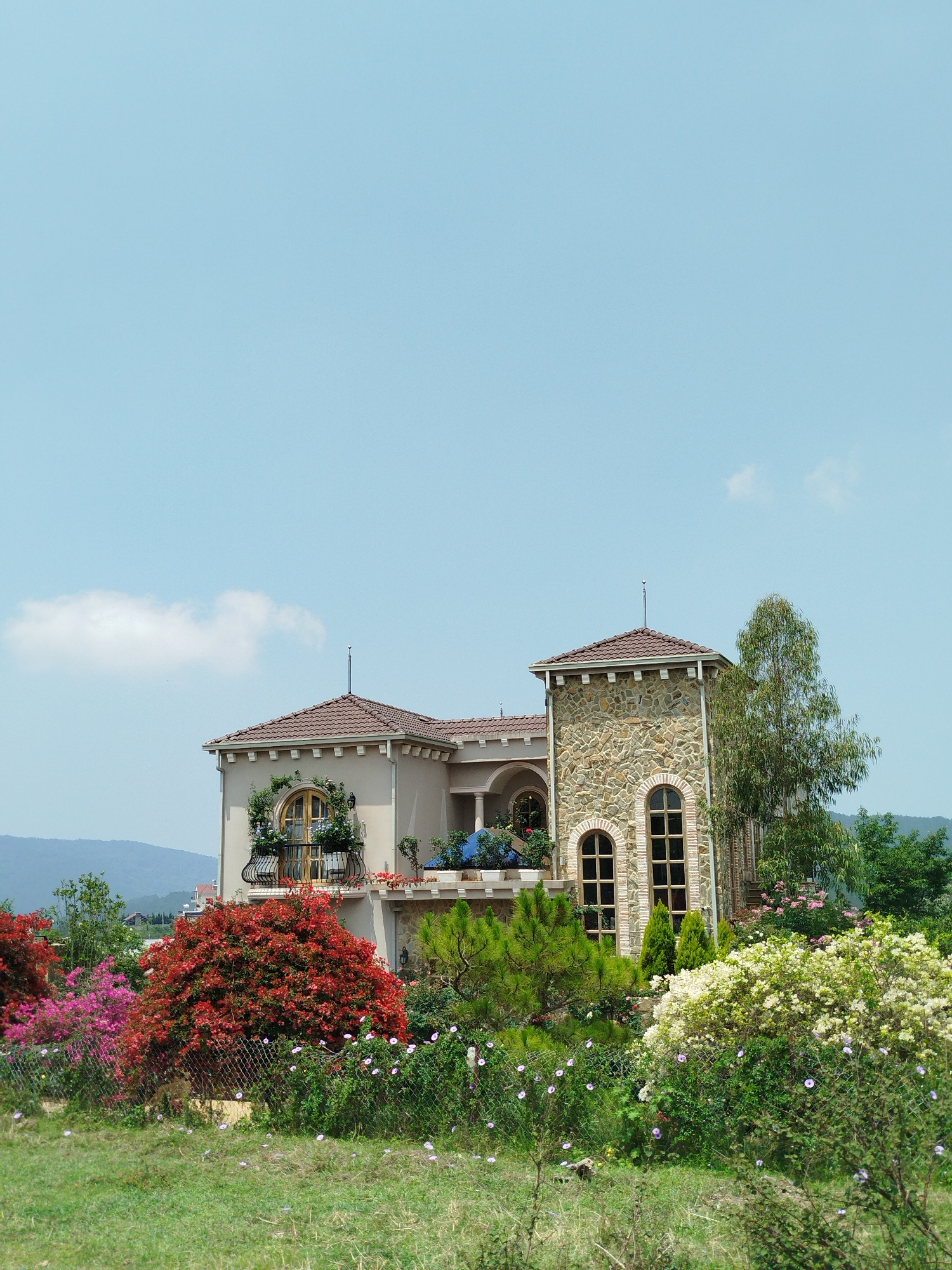 The image captures a two-story building, possibly a house, in what appears to be Lang Biang - Đà Lạt, Vietnam. The building features a beige exterior with a stone tower-like structure on the right side. Both the main structure and the tower have tiled roofs. A balcony extends from the second story, adorned with potted plants and flowers. The area surrounding the building is lush with colorful vegetation including red, pink, and white flowering bushes. A wire fence partially obscures the view of the foreground. In the background, mountains are visible under a clear blue sky with a single white cloud. No people are visible in the image.