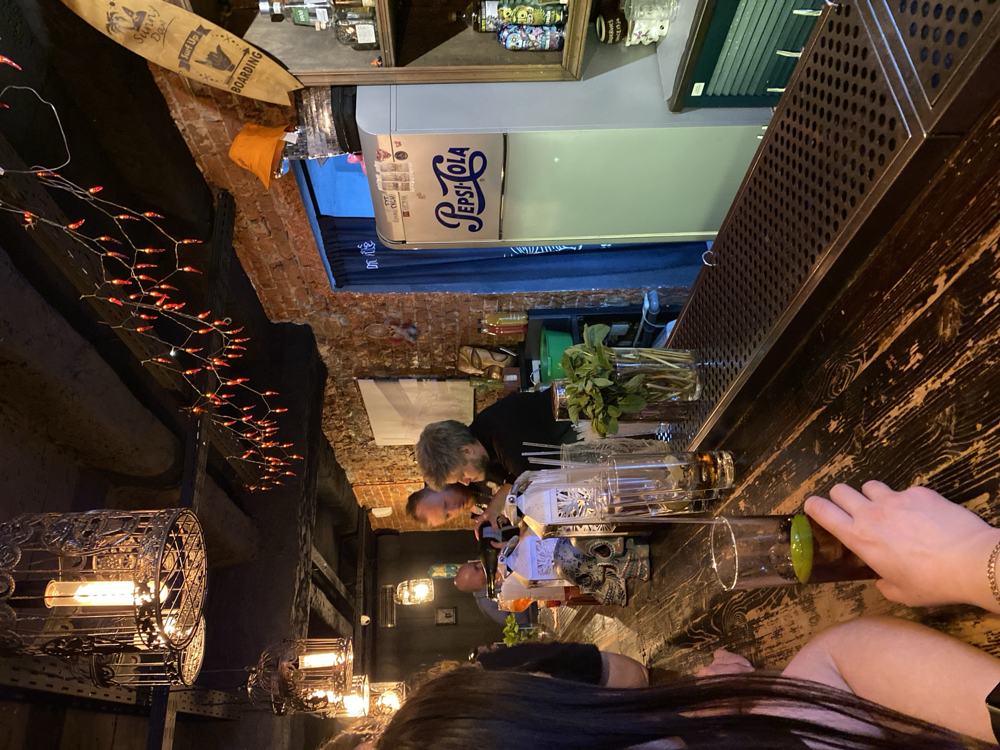 A dimly lit bar in Izhevsk, Russia, shows a bartender preparing drinks for patrons. The bar has a wooden counter, hanging decorative lamps, and exposed brick walls. A "Pepsi-Cola" branded refrigerator is visible. Patrons are seated at the bar, with arms and drinks visible on the counter.