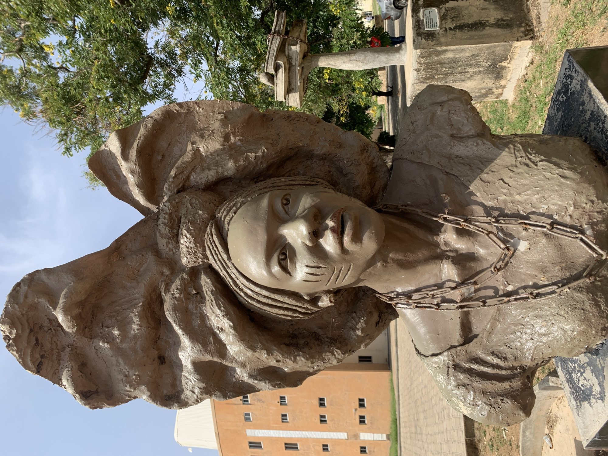 The image features a close-up of a weathered, earthen-toned statue of a woman. She is adorned with a large, sculpted head covering, and facial markings are visible on her cheeks. A chain rests around her neck. The background includes a green tree and an orange multi-story building. The image appears to be taken outdoors, likely in daylight. This is a statue of Moremi Ajasoro located in Jos, Nigeria.