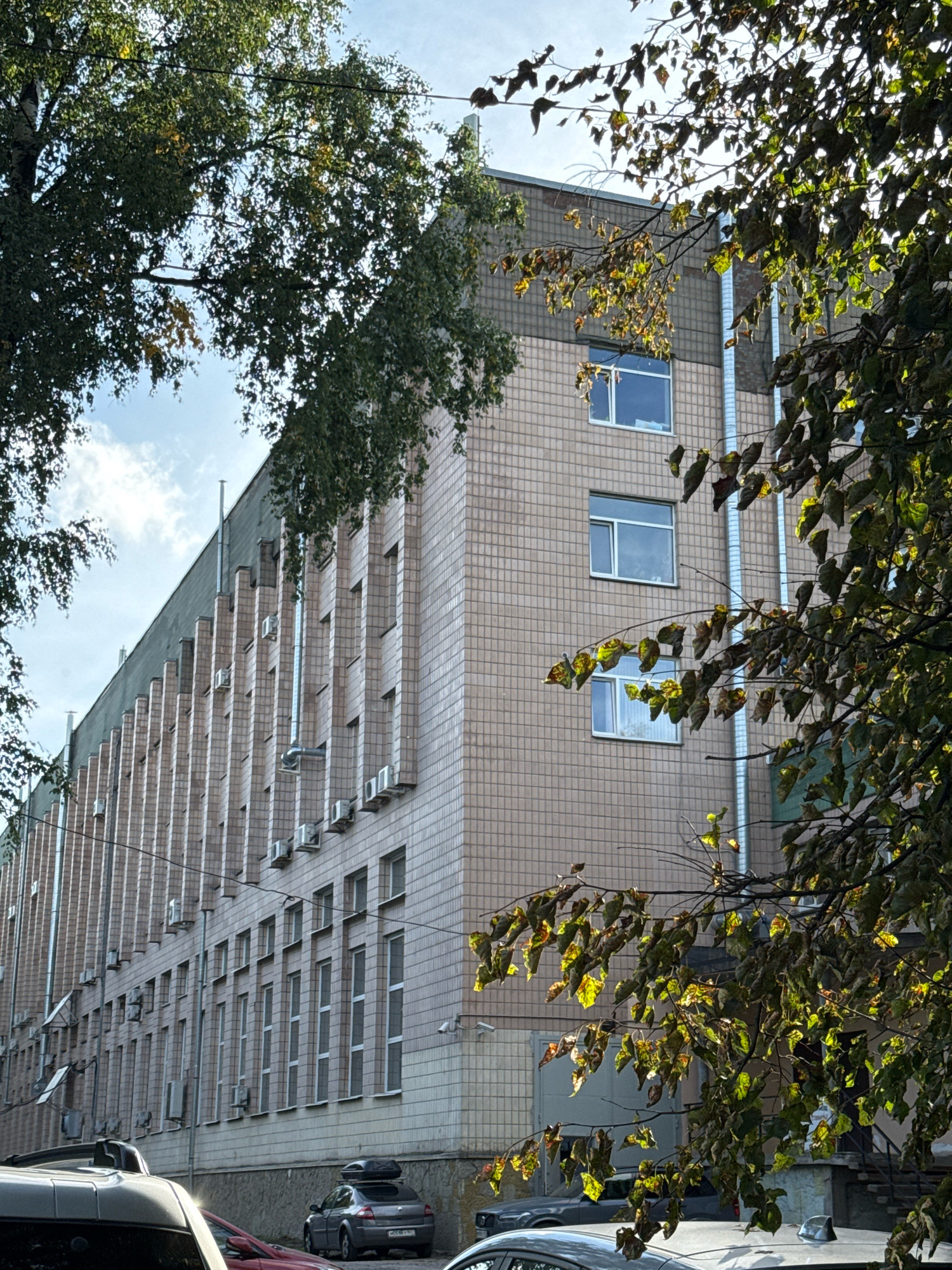 The image shows a multi-story building with a facade covered in light-colored square tiles. The building has a series of evenly spaced vertical elements that create a textured appearance. Numerous air conditioning units are mounted on the facade. Several windows are visible, some with white frames. Tall metal pipes run vertically along the building. In the foreground, the branches of a tree with green and dried leaves partially obscure the view of the building. In the street below the building, a gray car with a black roof box is parked. Another red car is partially visible behind it. The sky is visible with scattered clouds. The scene appears to be an urban environment, likely in Saint Petersburg, Russia, given the architectural style and the presence of trees commonly found in the city.