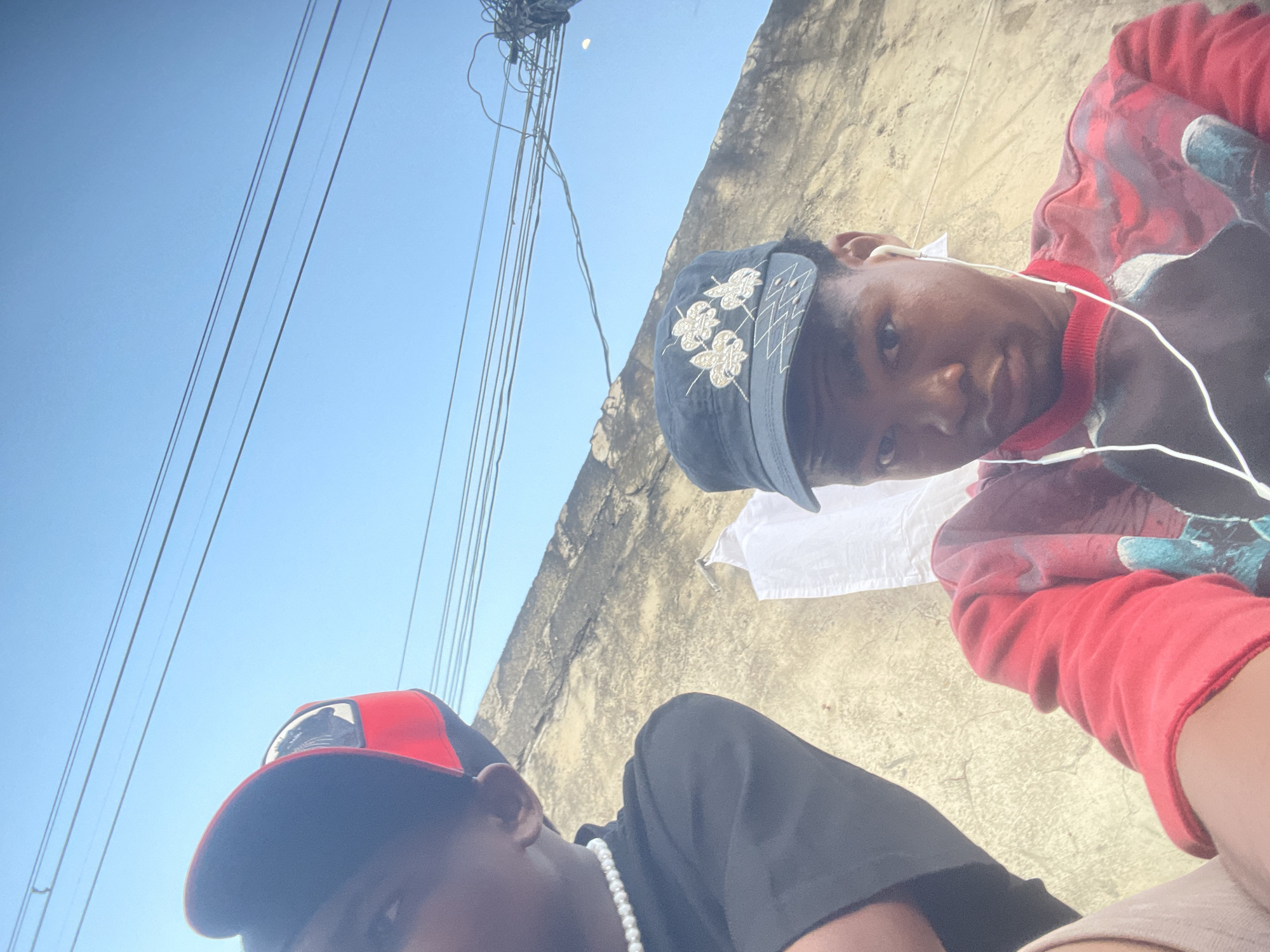 Two young men are pictured outdoors, with a rough, textured wall and a clear blue sky in the background. Power lines are visible against the sky. The man on the left, partially visible, wears a black t-shirt and a red and black baseball cap, along with a pearl necklace. The man on the right wears a dark blue cap with embroidered white designs, a red sweatshirt with a graphic print, and white earphones. He is looking directly at the camera. The setting appears to be an urban environment in Ado, Nigeria, possibly a residential or public area with exposed building materials and overhead utilities.