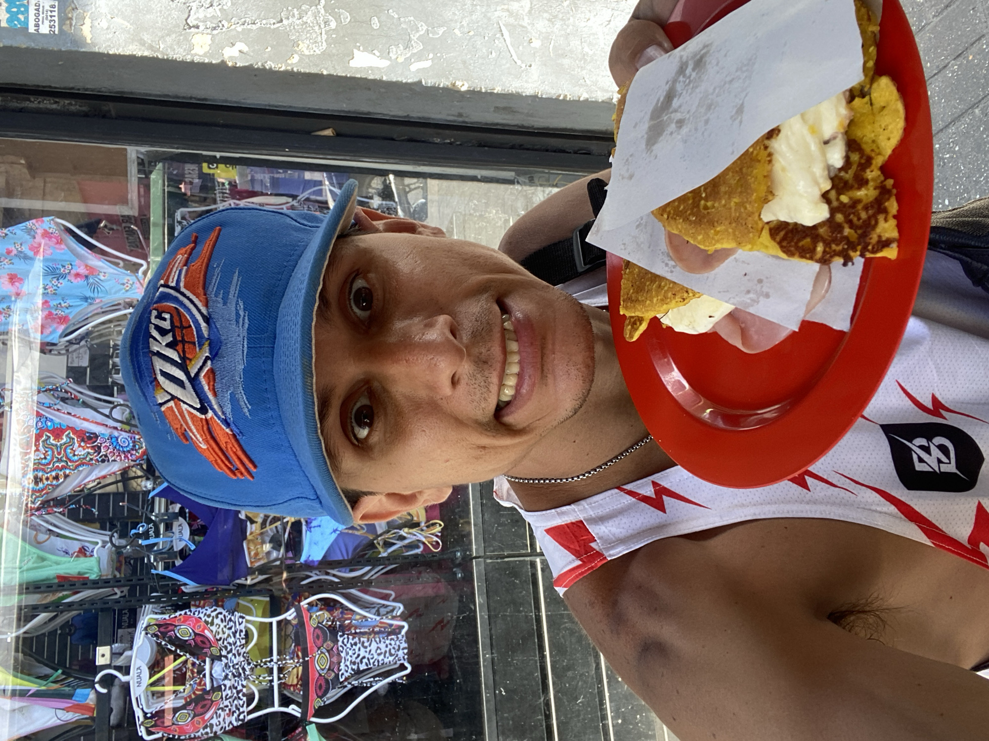 A male individual is shown in a close-up, eye-level selfie, holding a red plate containing two cachapas filled with white cheese and ham, partially covered by white paper. He has light brown skin, dark eyes, and an open-mouthed, wide-eyed expression. He wears a blue baseball cap with an "OKC Thunder" logo and a white athletic singlet with red lightning bolt designs and a black stylized logo on the left chest area. A silver chain is visible around his neck, and a bruise is present on his left shoulder. The background features a store window displaying various women's swimwear, with "NUALA" branding visible on some hangers. To the right of the individual, a weathered light-colored wall or pillar with peeling paint exhibits a partially visible blue sign displaying the text "ABOGADO" and "253118". The presence of the "NUALA" brand and the "ABOGADO" sign with a local phone number, combined with the street food (cachapas), indicates a commercial street environment in the city of Caracas, Venezuela.
