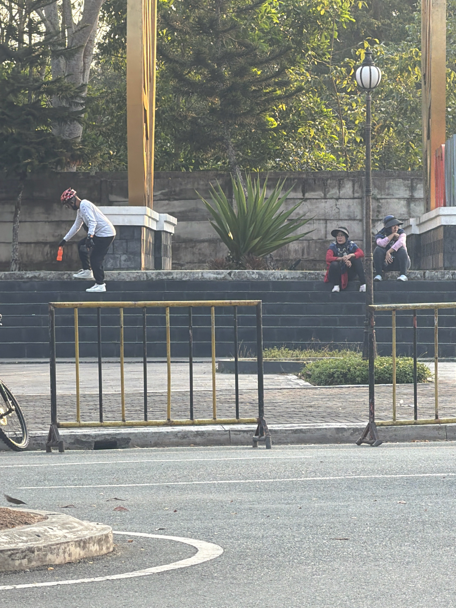 A cyclist wearing a white long-sleeved shirt, black pants, white shoes, and a red and white helmet is on a set of stairs in Balikpapan, Indonesia, holding an orange bottle. Two women are seated on the same stairs further back. One woman wears a red and blue jacket and black pants, while the other wears a pink long-sleeved shirt and black pants. Both women wear hats. A lamppost stands behind them. In the foreground, a yellow and black metal barrier runs horizontally across the image, and a bicycle wheel is visible on the left side. The background consists of trees and a large, golden, rectangular structure. The scene appears to be set in a park or public area with paved pathways and dense foliage.