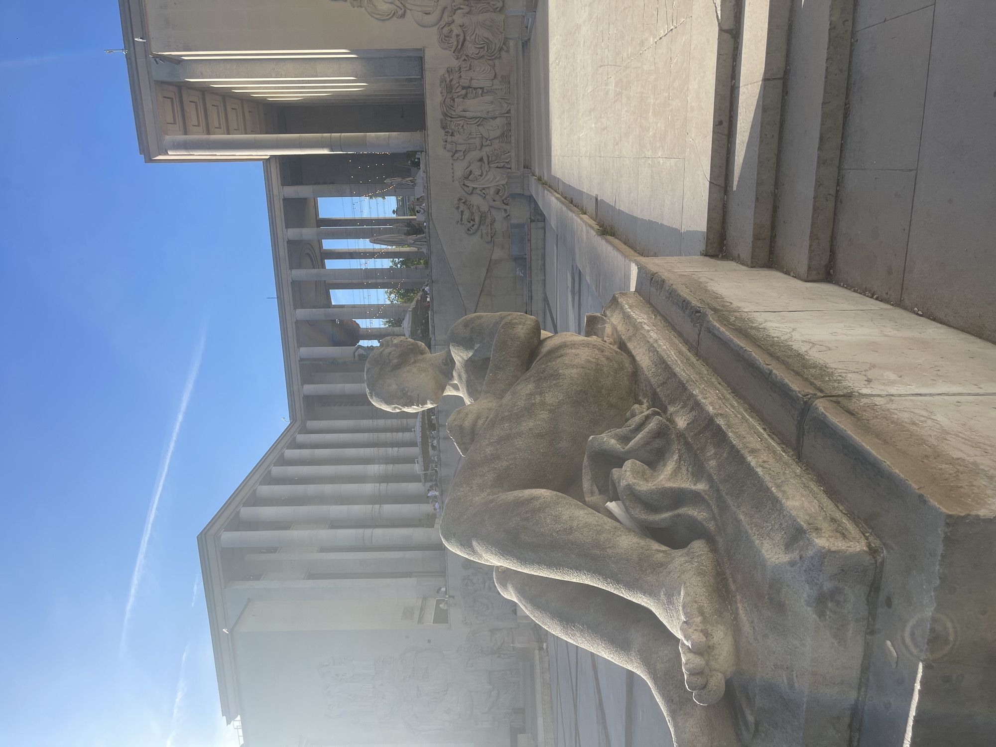 The image shows a sunlit outdoor scene in front of a large, neoclassical building with a colonnade. In the foreground, a stone sculpture of a reclining nude figure is prominently displayed on a raised platform. The sculpture depicts a person with legs crossed, holding an object in their hands. The building behind the sculpture features a long, symmetrical facade with numerous columns and decorative reliefs along its base. Steps lead up to the building's entrance. The sky is clear blue with a faint contrail. The overall impression is one of grandeur and public space. This scene is located at the Palais de la Porte Dorée in Paris, France, a building that now houses the National Museum of Immigration History.